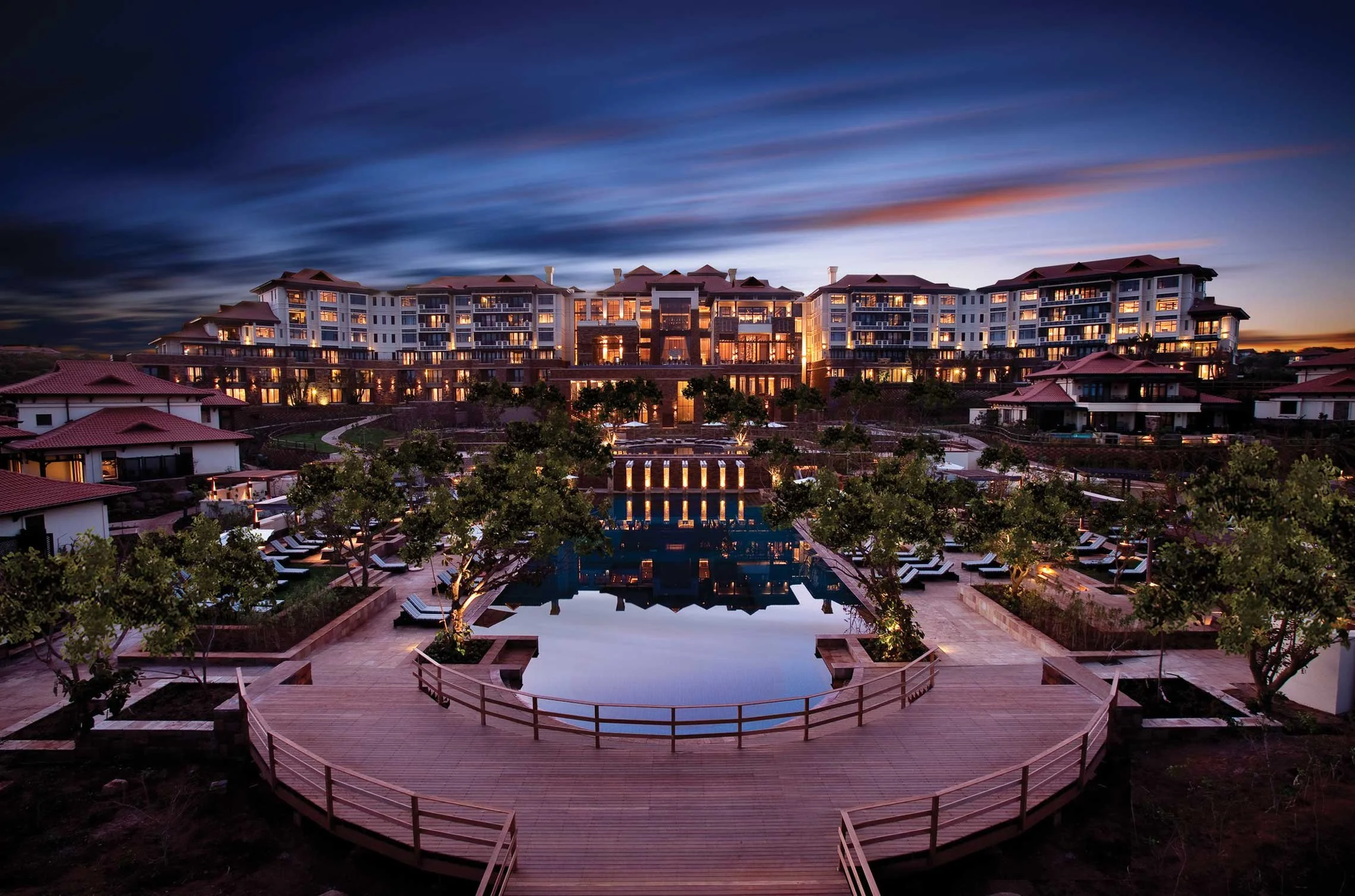 Luxury multi-story residential building at dusk with illuminated windows, a reflecting pool in front, surrounding trees, and a circular wooden walkway with benches.