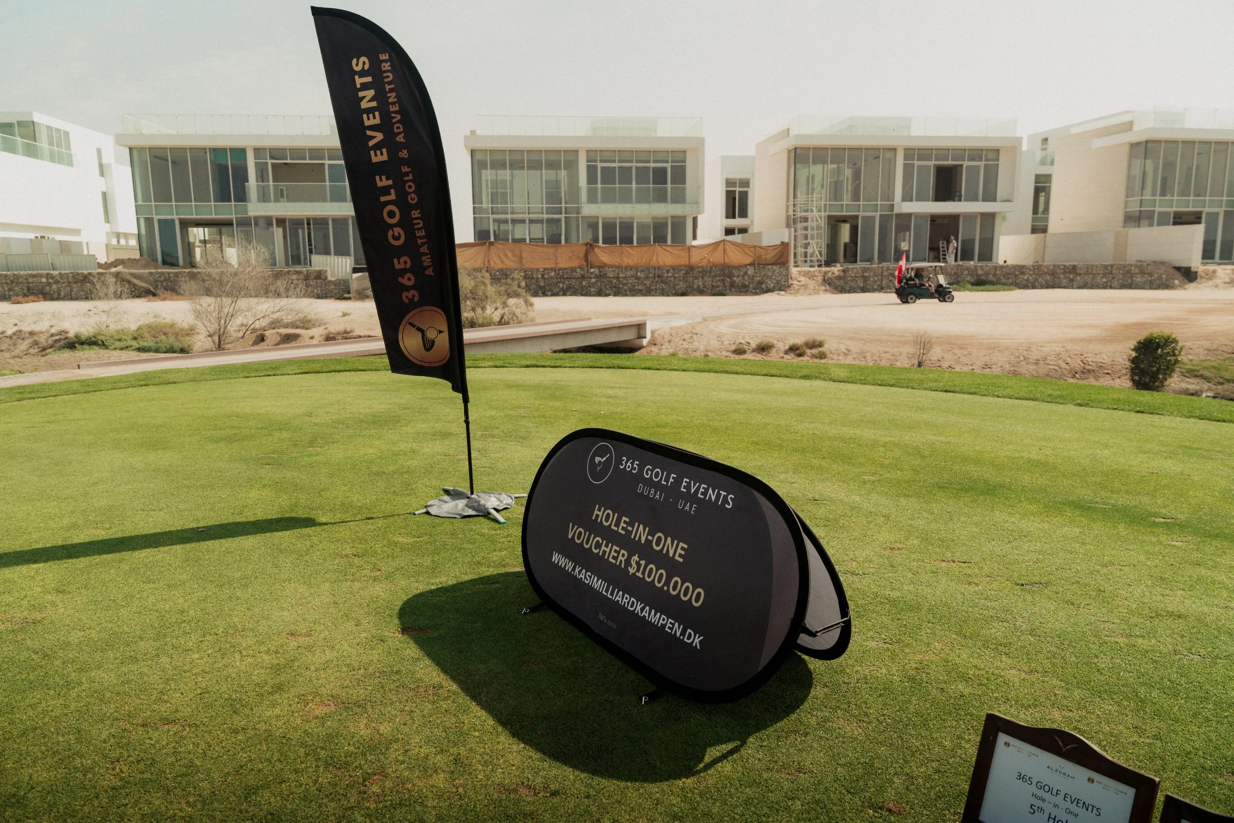 Signage at a golf course promoting a hole-in-one contest with a $100,000 voucher, surrounded by neatly maintained grass and modern residential buildings in the background.