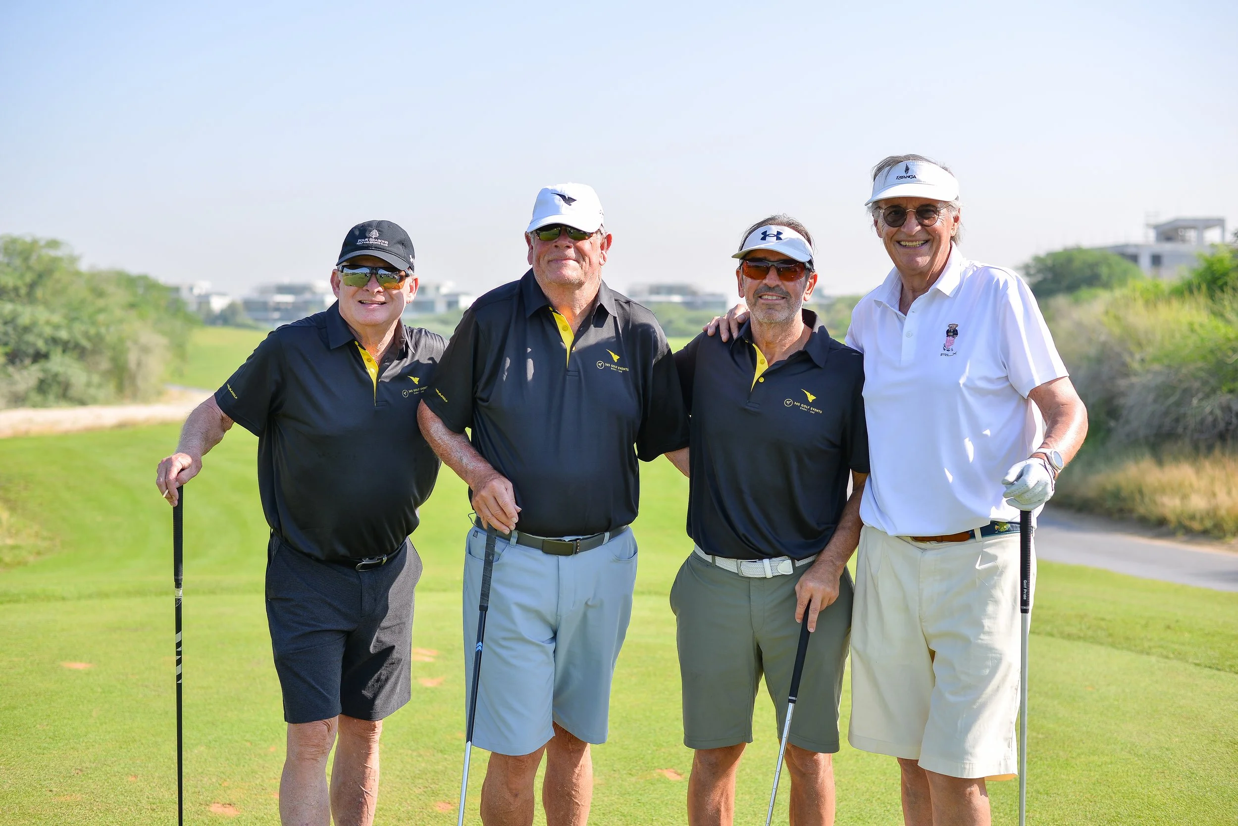 Four seniors smiling and posing with golf clubs on a golf course, dressed in casual golf attire with sunglasses and hats.