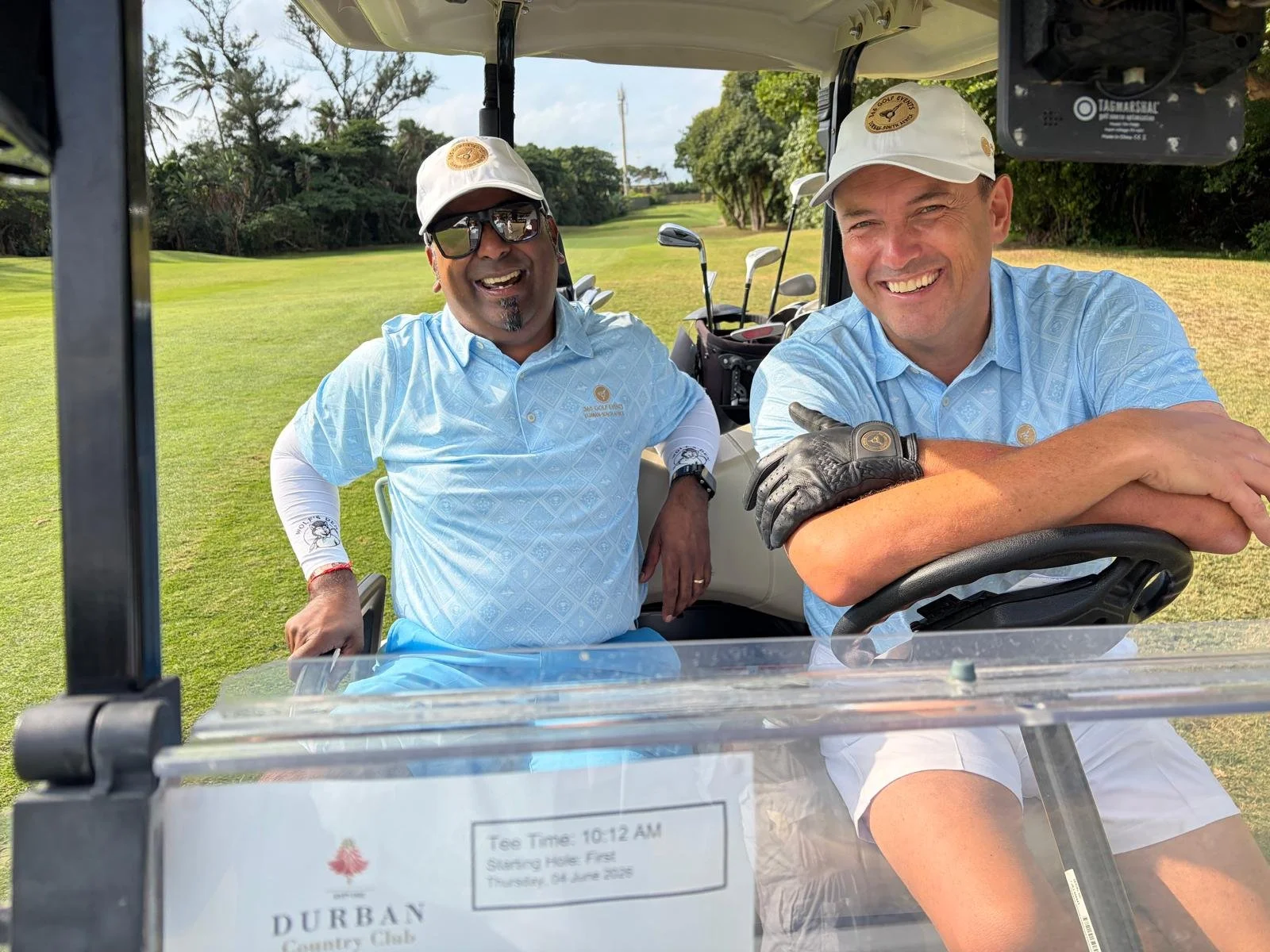 Two men smiling and sitting in a golf cart on a golf course, with golf clubs in the background and lush green trees around.