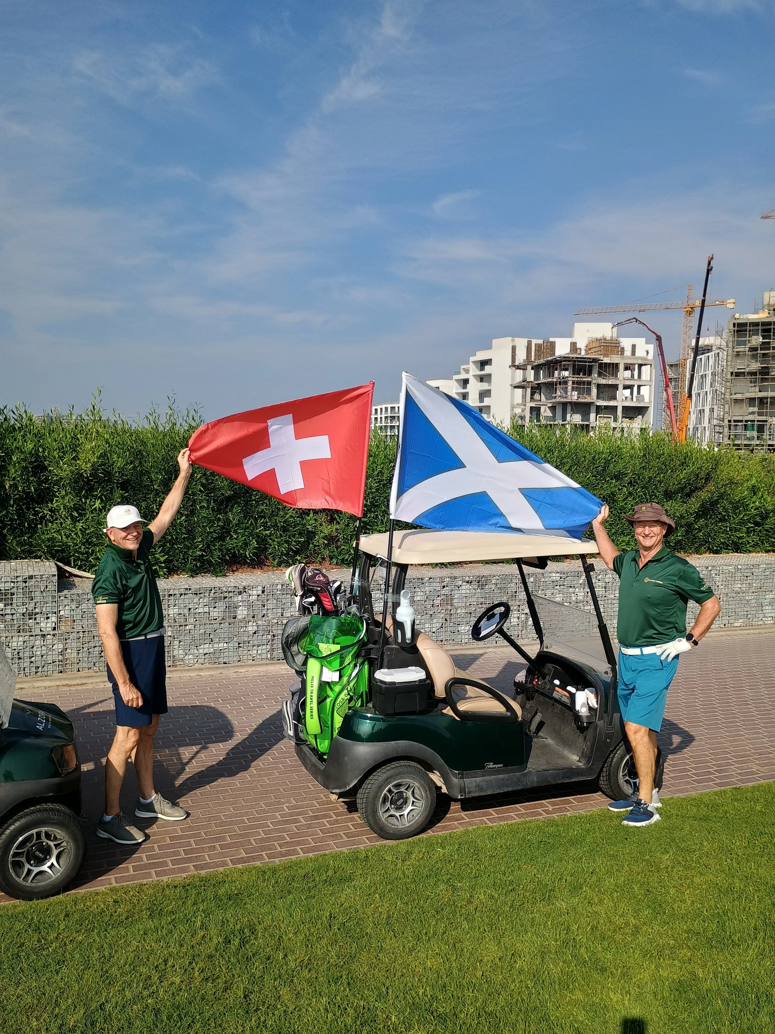 Two men standing beside a golf cart with flags of Switzerland and Scotland, on a golf course with buildings and construction cranes in the background.