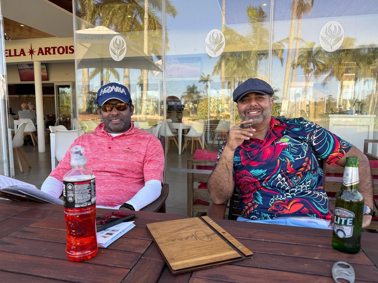 Two men sit at an outdoor restaurant table on a sunny day. The man on the left wears a pink shirt, sunglasses, and a blue hat, while the man on the right wears a colorful Hawaiian shirt, a cap, and smokes a cigar. The table has a bottle of beer, a bo