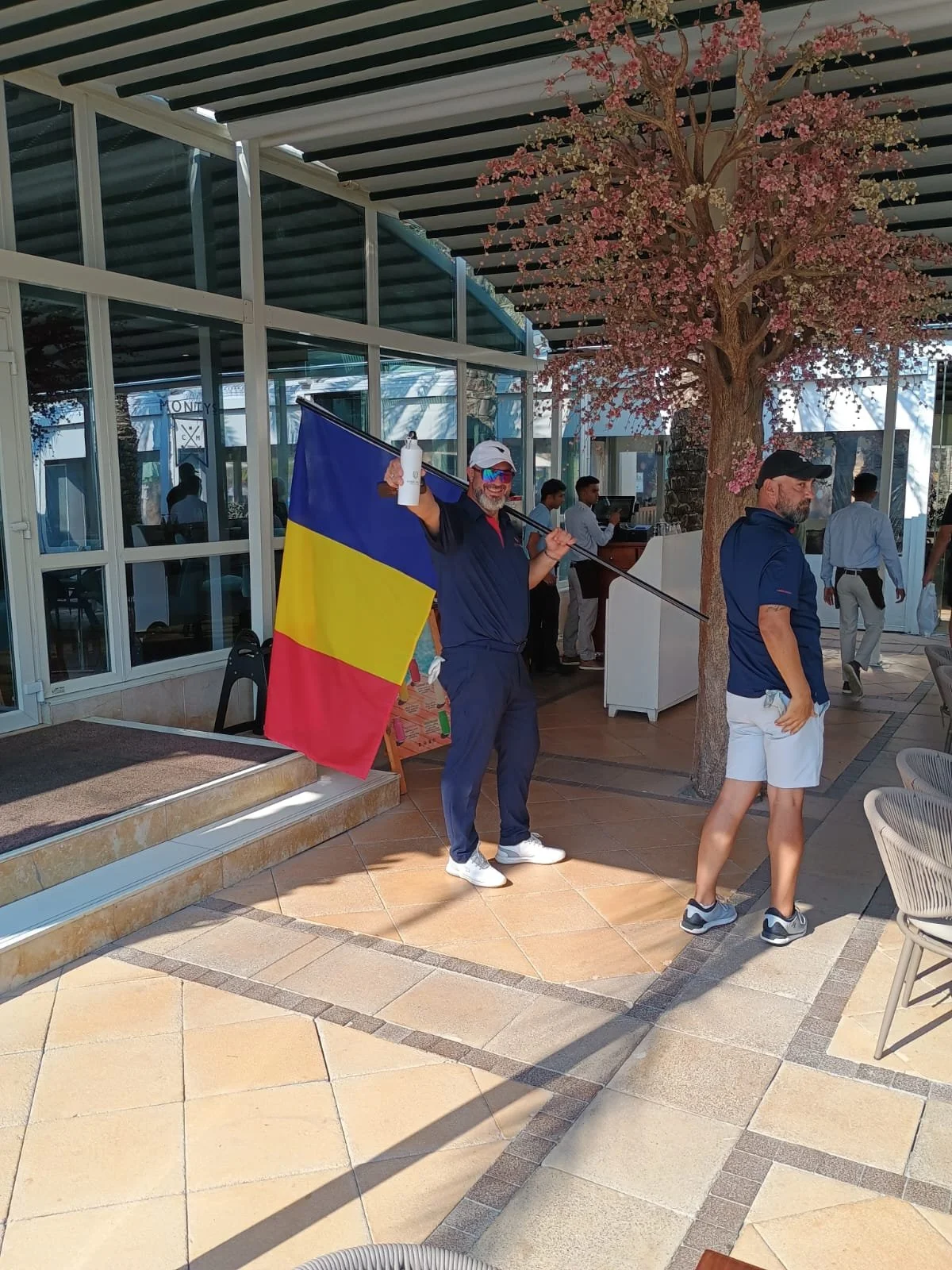 Man holding a flag with the colors blue, yellow, and red, standing outdoors near a pink flowering tree, with other people in the background.