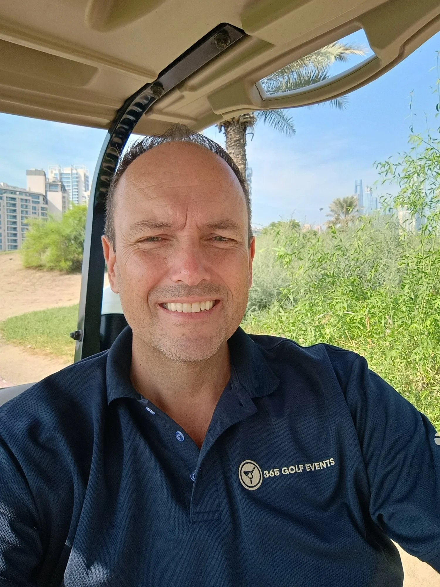 A man smiling sitting in a golf cart outdoors with palm trees, bushes, and city buildings in the background, wearing a navy blue polo shirt with a logo that says "365 GOLF EVENTS."