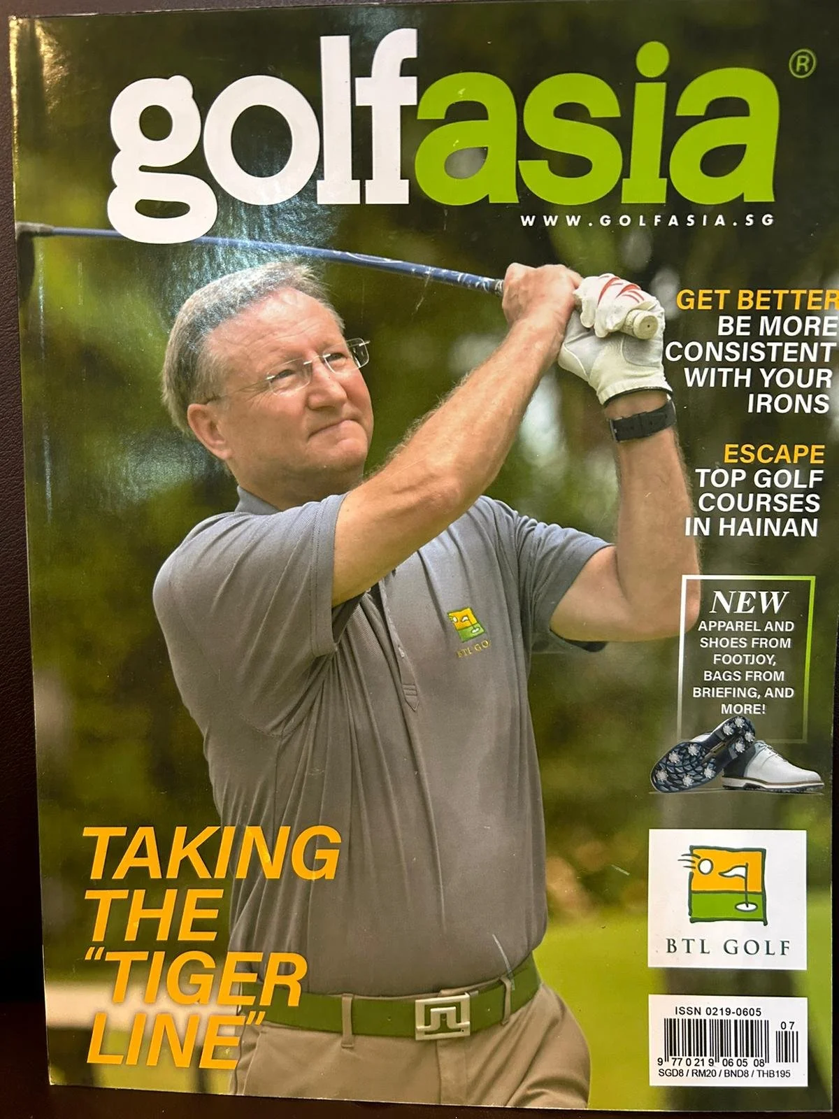 A man in a gray polo shirt and glasses is taking a golf swing on a golf course, with trees in the background. The magazine cover features the title 'golf asia' and various headlines about golf tips, courses, and products.