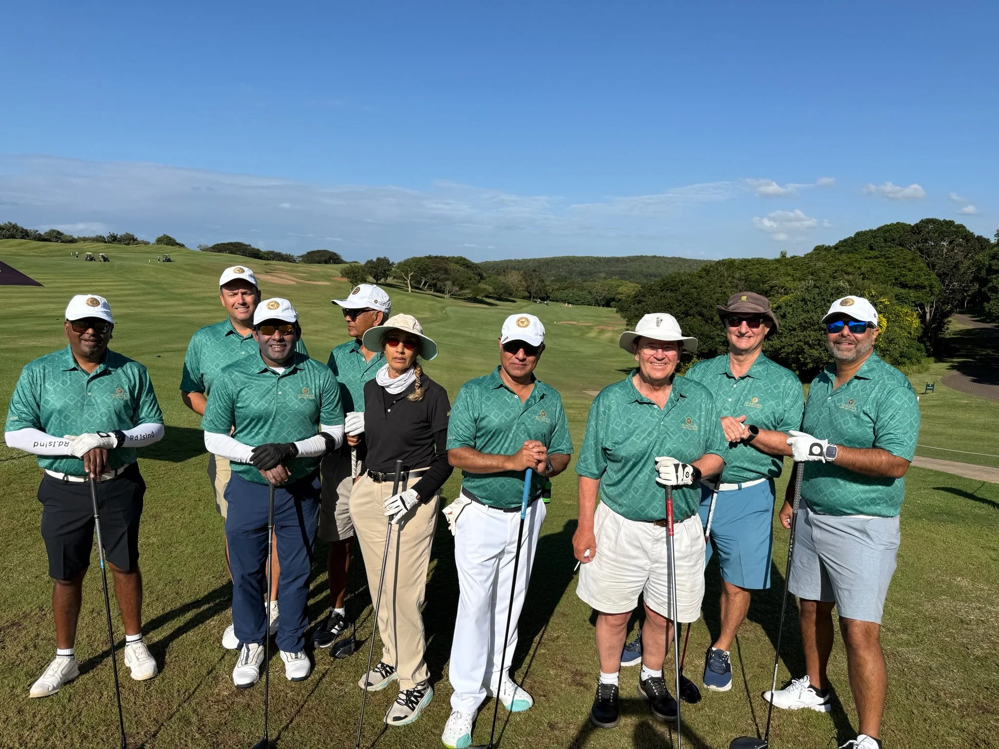 A group of nine people standing on a golf course with golf clubs, wearing matching teal shirts and hats, under a blue sky with some clouds, surrounded by green trees and rolling hills.