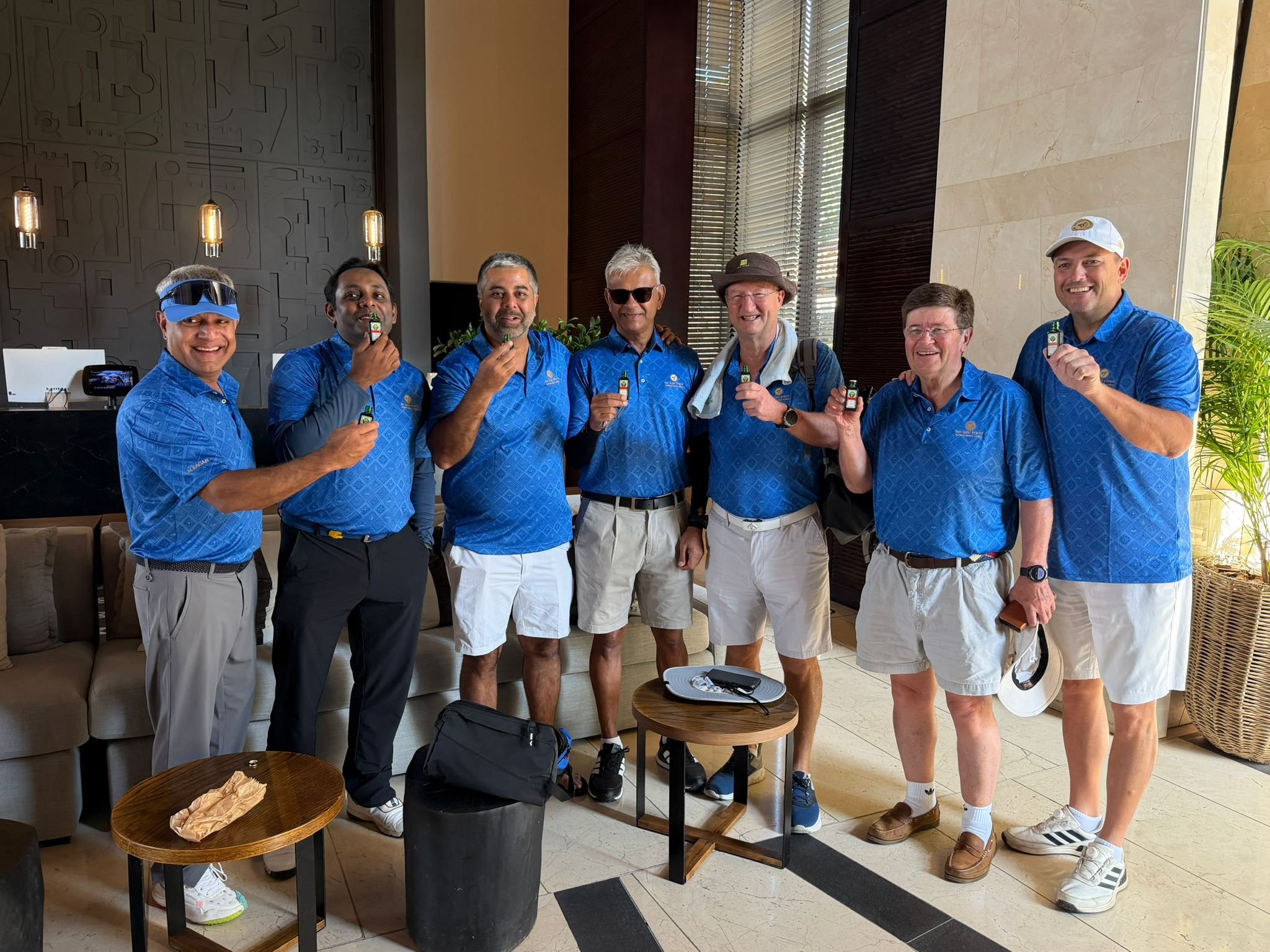 A group of seven men posing for a photo indoors, all wearing blue shirts, some with golf caps or hats, holding small bottles, smiling, with a table and plants visible in the background.
