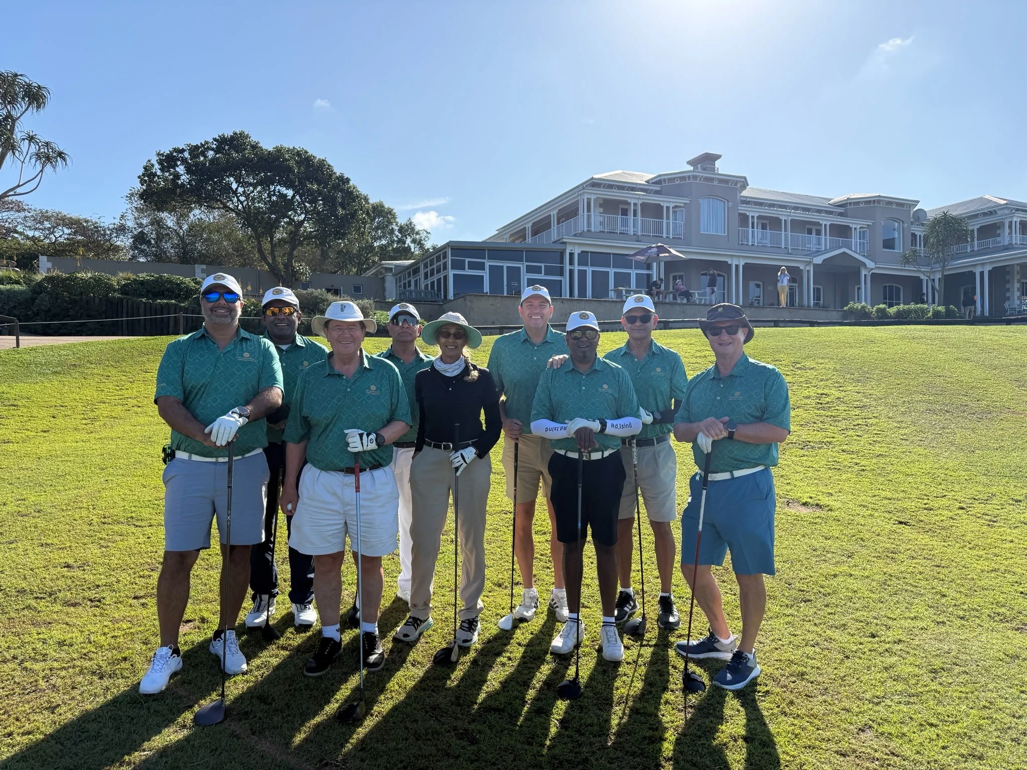 Group of nine people standing on a golf course holding golf clubs, wearing matching teal shirts, white gloves, and sun hats, with a large house and trees in the background under a sunny sky.