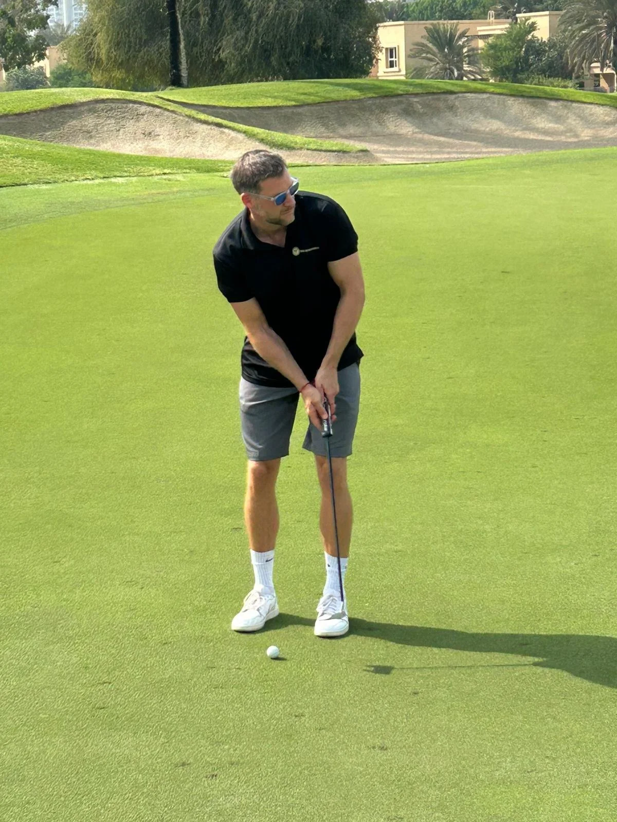 A man standing on a golf course, preparing to putt a golf ball, wearing sunglasses, a black shirt, gray shorts, white sneakers, and white socks, with sand bunkers and palm trees in the background.