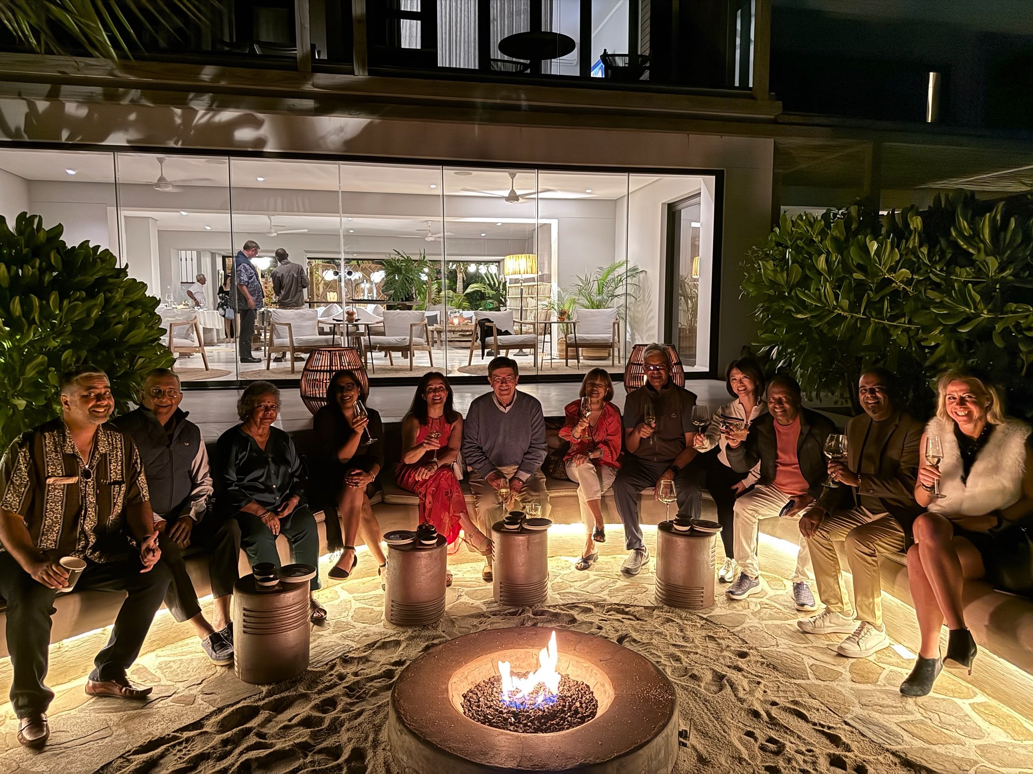 A group of people sitting and smiling around a fire pit at night with a large glass window showing an indoor room filled with plants and furniture behind them.