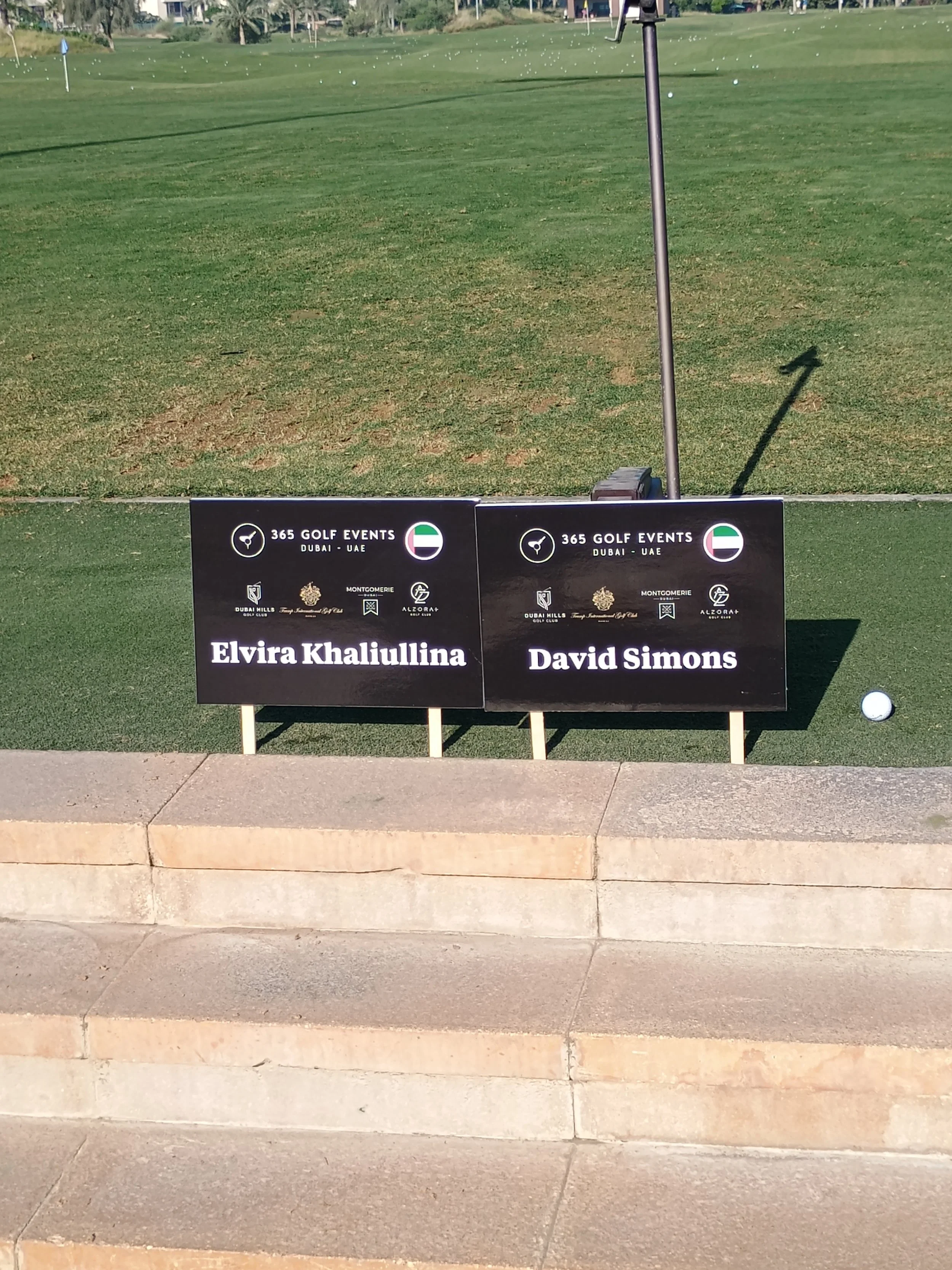Leaderboard signs for a golf event at Dubai course, displaying the names of Elvira Khaliullina and David Simons, with golf balls and a golf tee nearby.