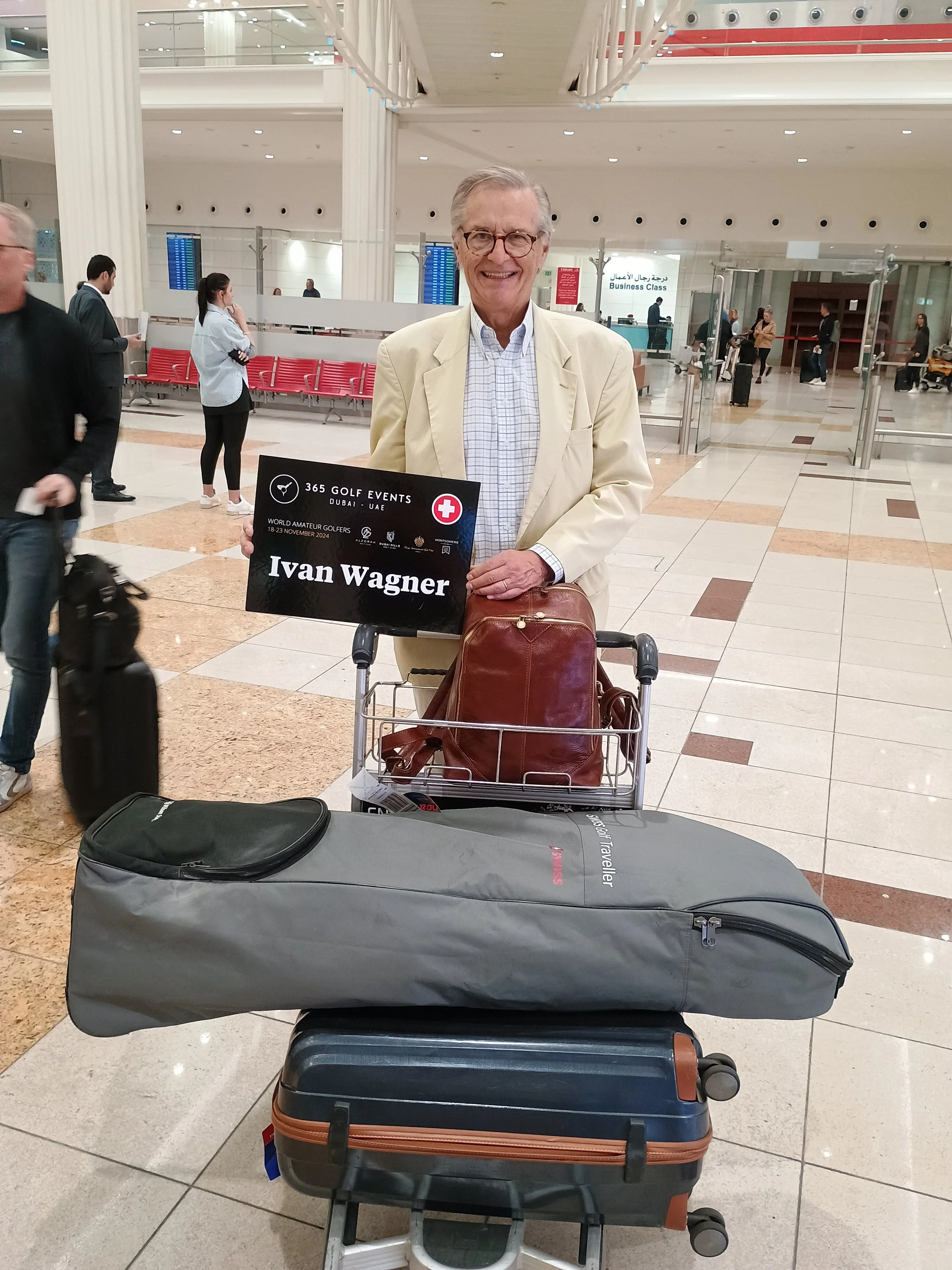 An elderly man in a cream-colored jacket and checkered shirt smiles at the camera while standing in an airport terminal with a luggage cart. The cart has a large gray suitcase, a black toiletry bag, and a brown leather bag. He is holding a sign that reads 'Ivan Wagner' with a logo indicating it is for a golf event in Dubai, UAE. Other travelers are in the background, and the airport has a modern design with white columns and a high ceiling.
