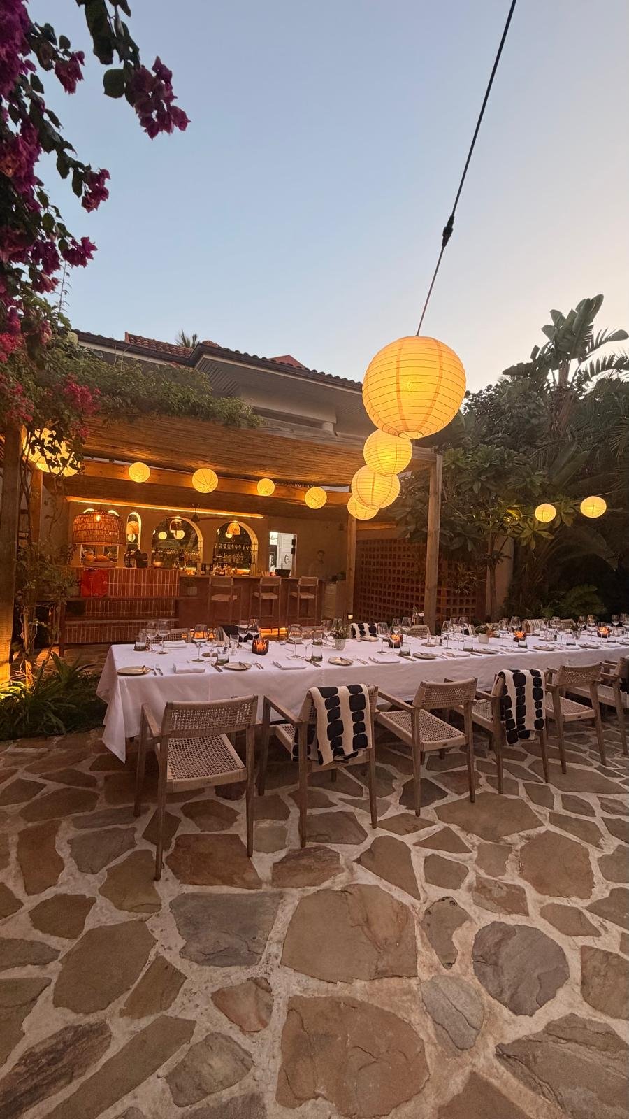An outdoor dining area set for a meal with a long table covered with a white tablecloth, decorated with glasses, plates, and candles. There are chairs around the table, some with black and white striped towels. Hanging yellow paper lanterns illuminat