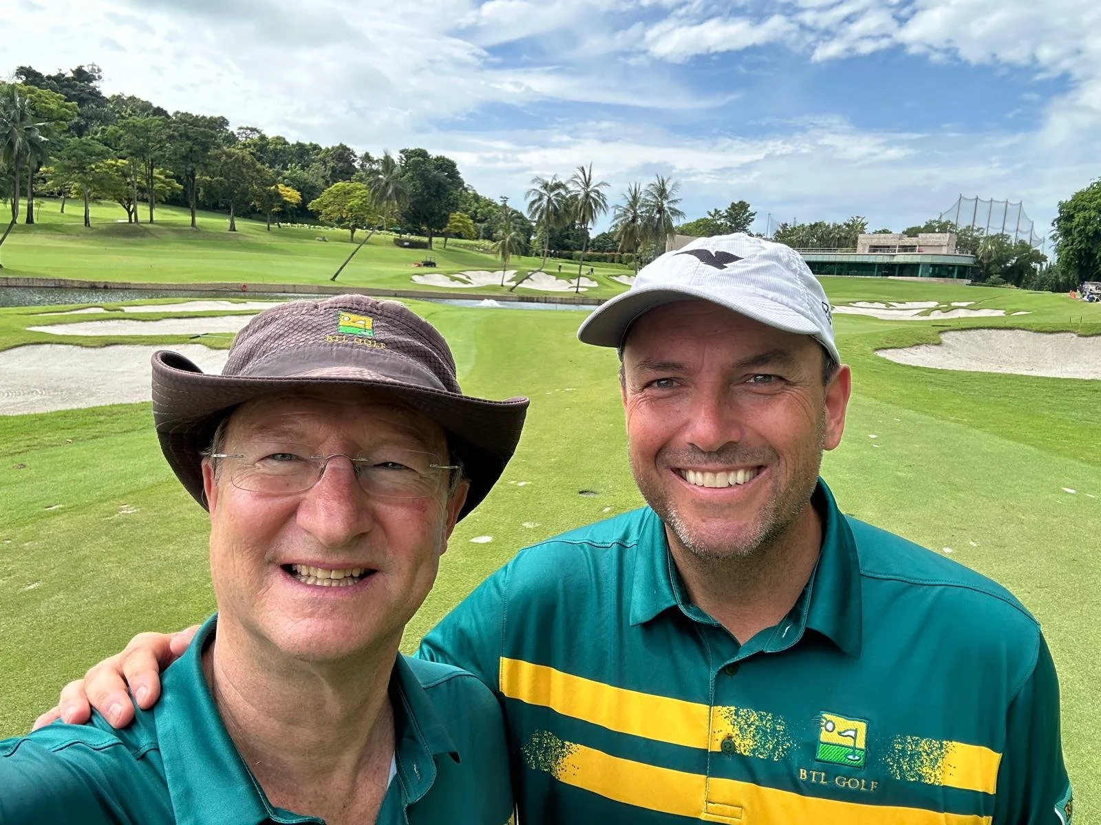 Two men smiling for a selfie on a golf course with green grass, sand bunkers, palm trees, and a building in the background.