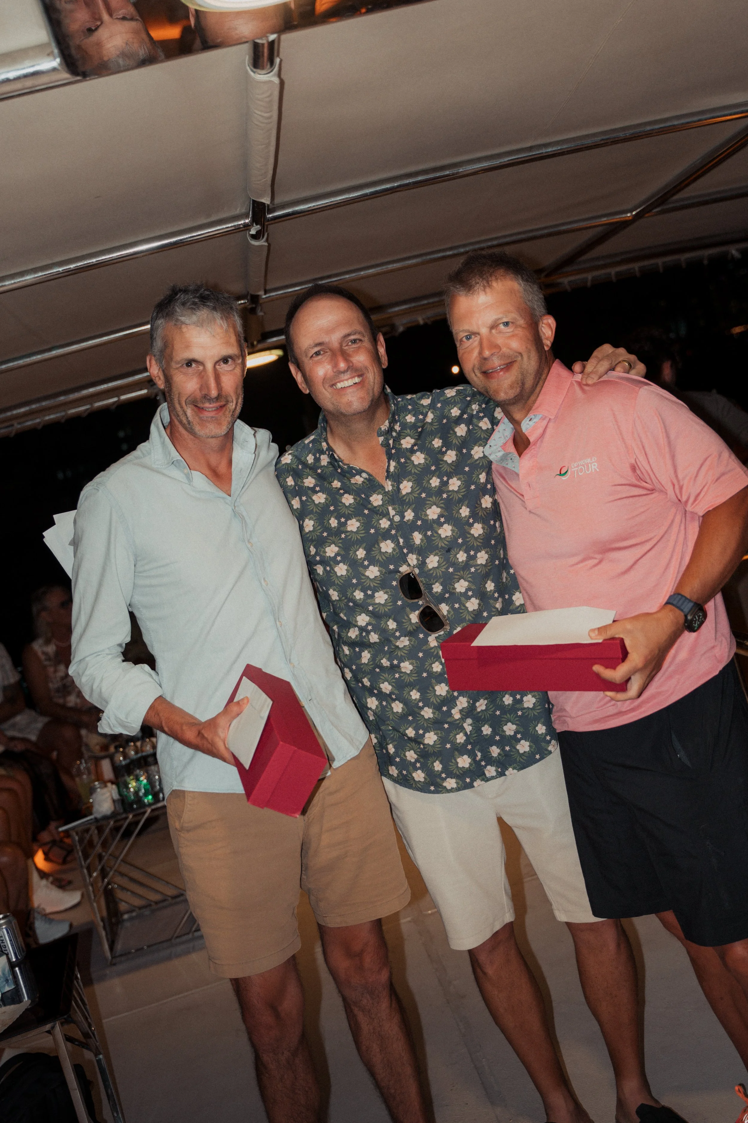 Three men standing together smiling, each holding a red gift box, at an outdoor evening event with a tent canopy overhead.