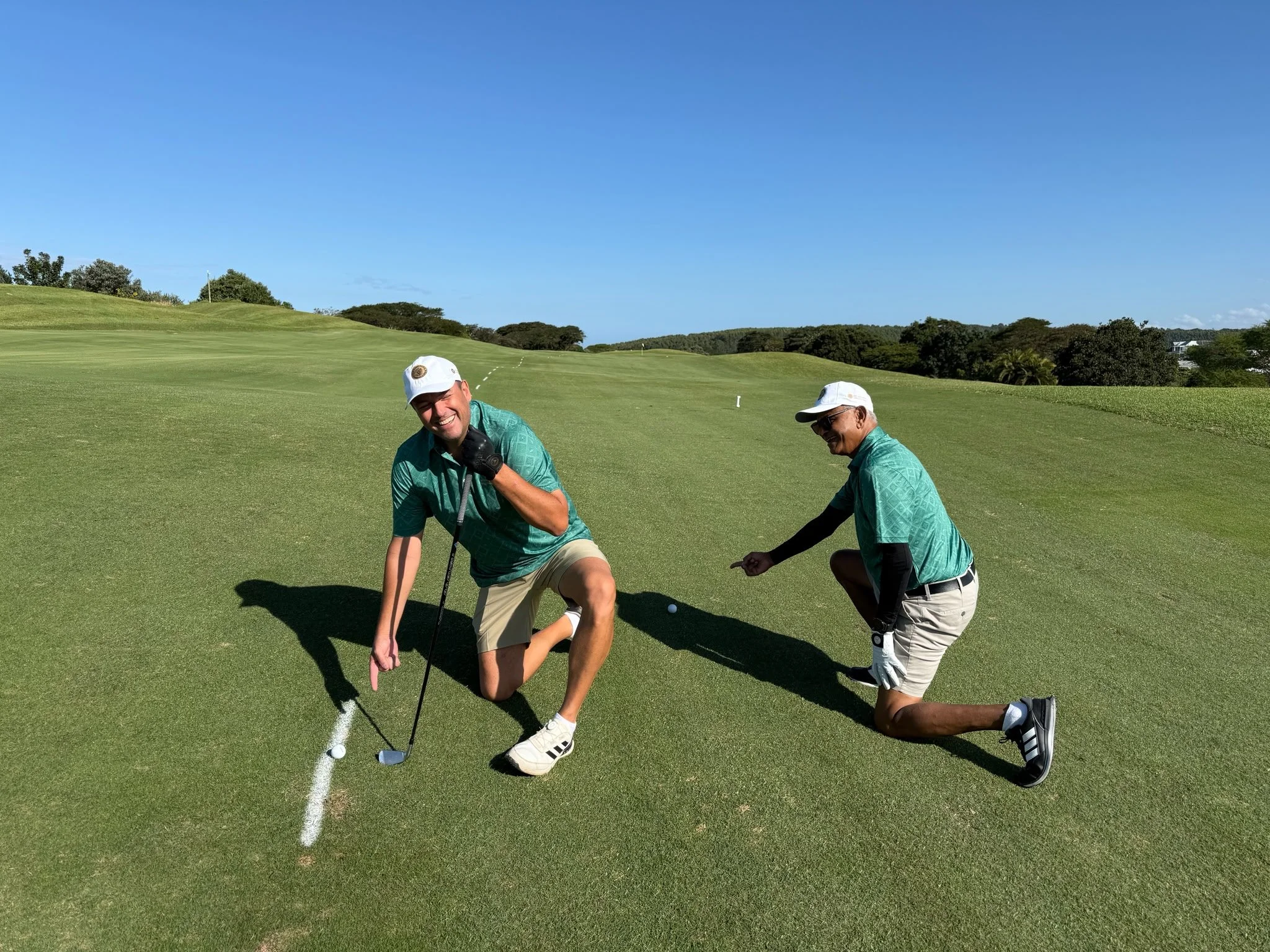 Two men on a golf course are kneeling and smiling at the camera, one holding a golf club, with a clear blue sky and green landscape in the background.