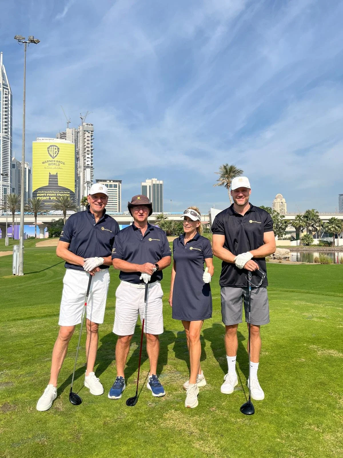 Four people standing on a golf course with city buildings in the background, holding golf clubs, dressed in golf attire, smiling, and posing for a photo.