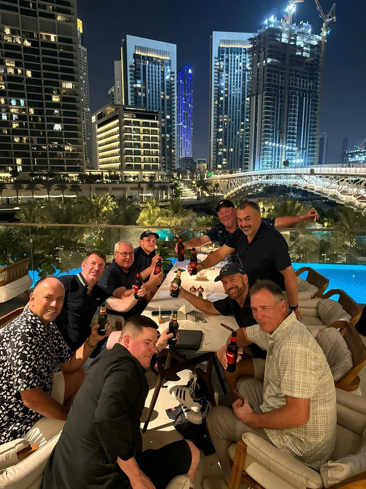 A group of men sitting around a table outdoors at night, with a city skyline and tall buildings illuminated in the background. They are holding bottles of beer and smiling.