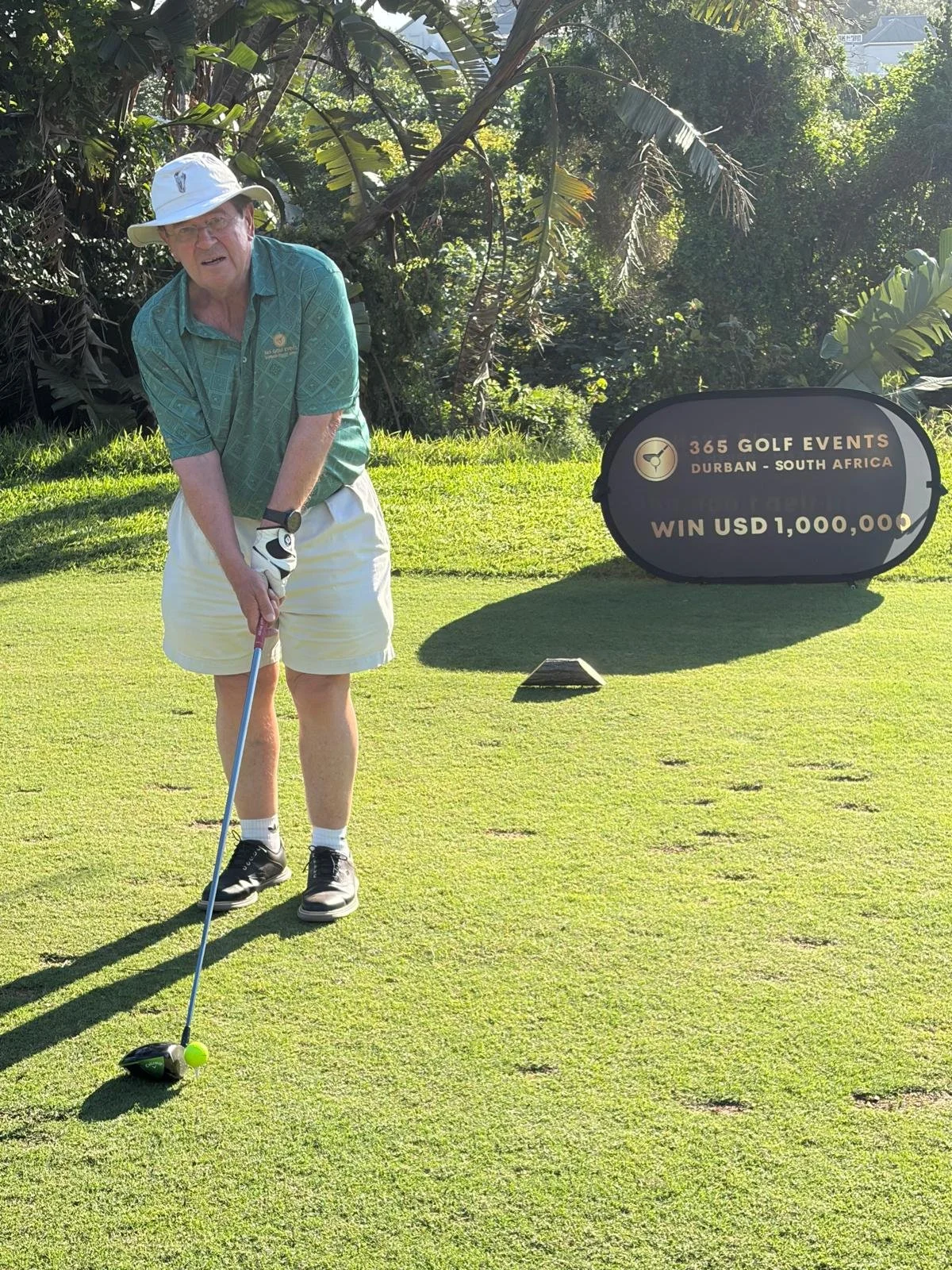 An elderly man playing golf on a golf course, wearing a light-colored hat, teal shirt, white shorts, and black shoes, preparing to hit the golf ball with a driver club. There is a large sign in the background that reads "365 GOLF EVENTS, Durban - Sou