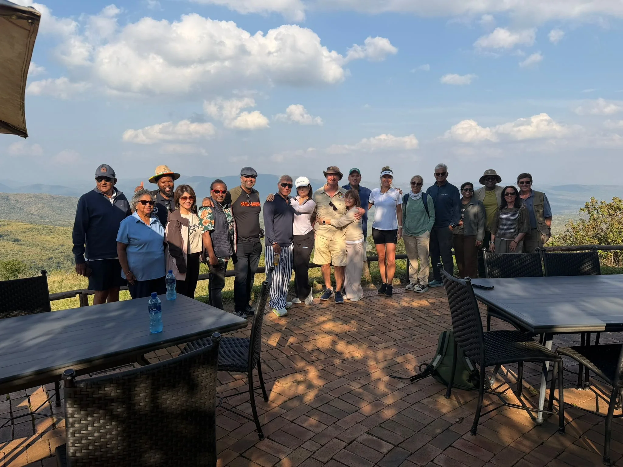 Group of 15 people standing outdoors on a sunny day with a scenic mountain view in the background, some wearing sunglasses and hats, posing for a photo.