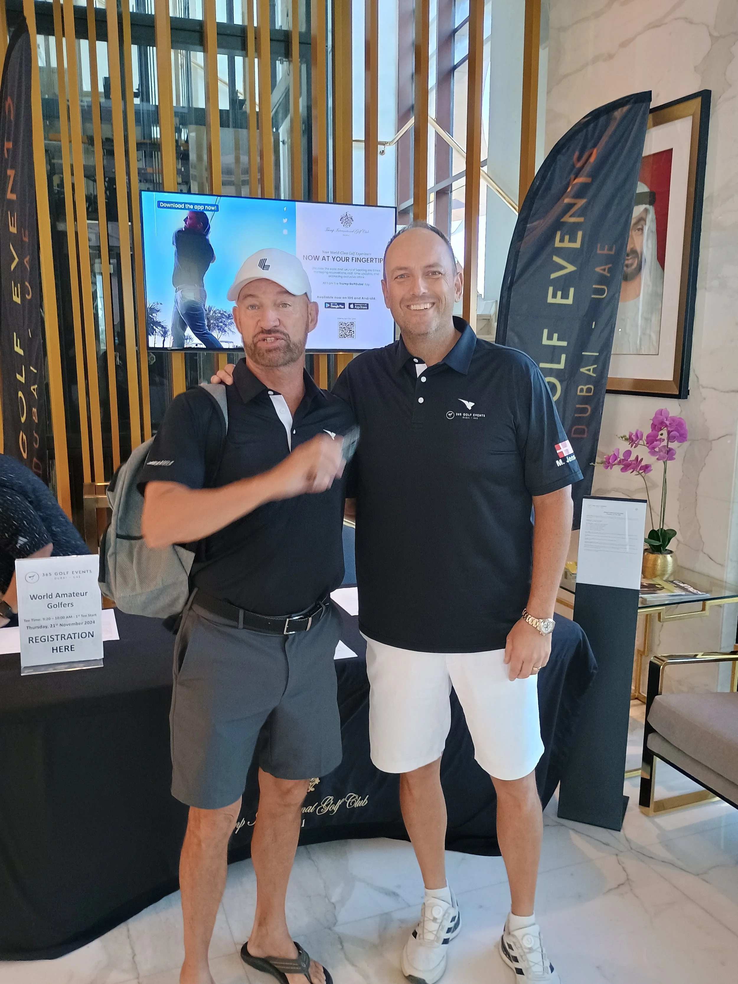 Two men in golf attire standing indoors, smiling for a photo. Behind them, a television screen displays a golfer and promotional content. There is a registration desk for a golf event with signs, and flags with 'Golf Events Dubai' written on them. The setting appears to be a hotel lobby or a conference area.