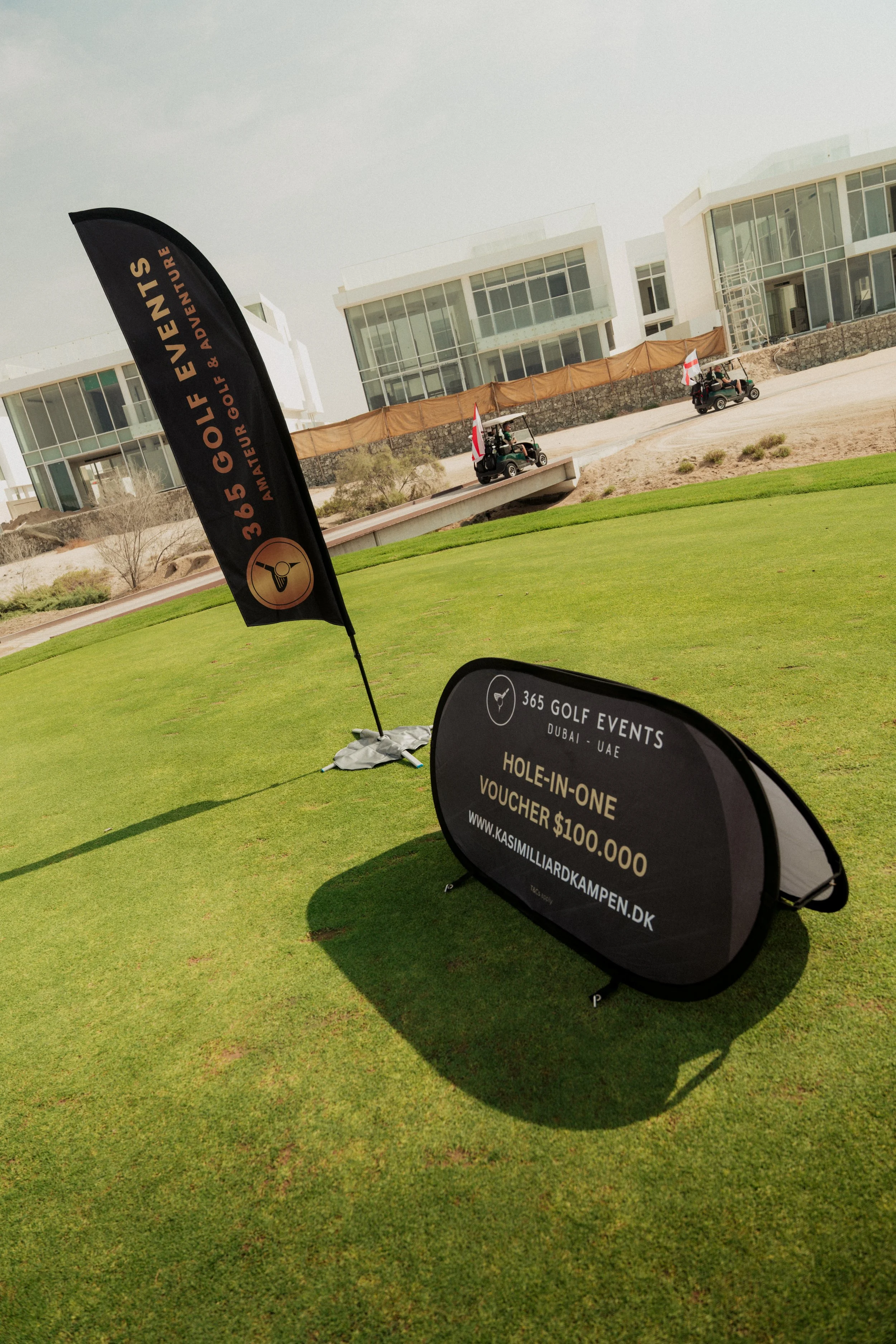 Sign promoting 365 Golf Events Dubai-UAE offering a hole-in-one voucher worth $100,000, with two golf carts and modern buildings in the background.