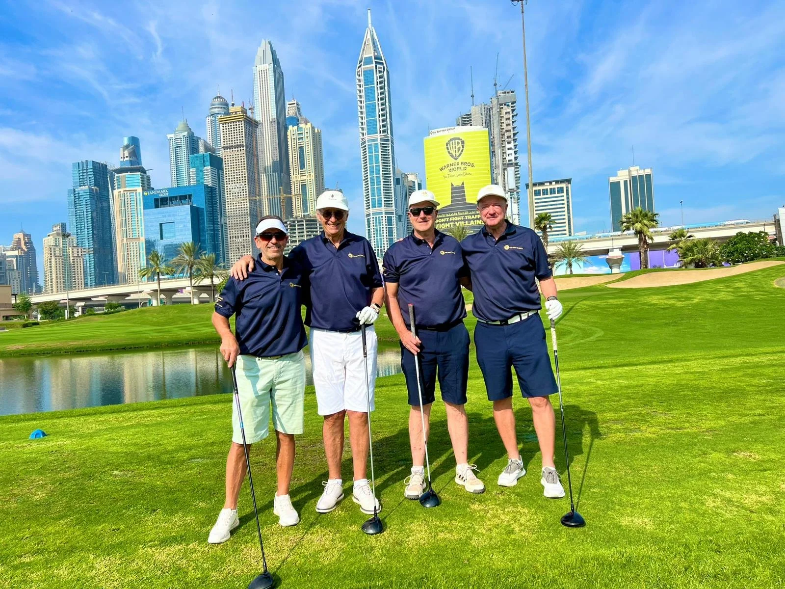 Four men in navy blue polo shirts and shorts standing together on a golf course with a city skyline in the background.