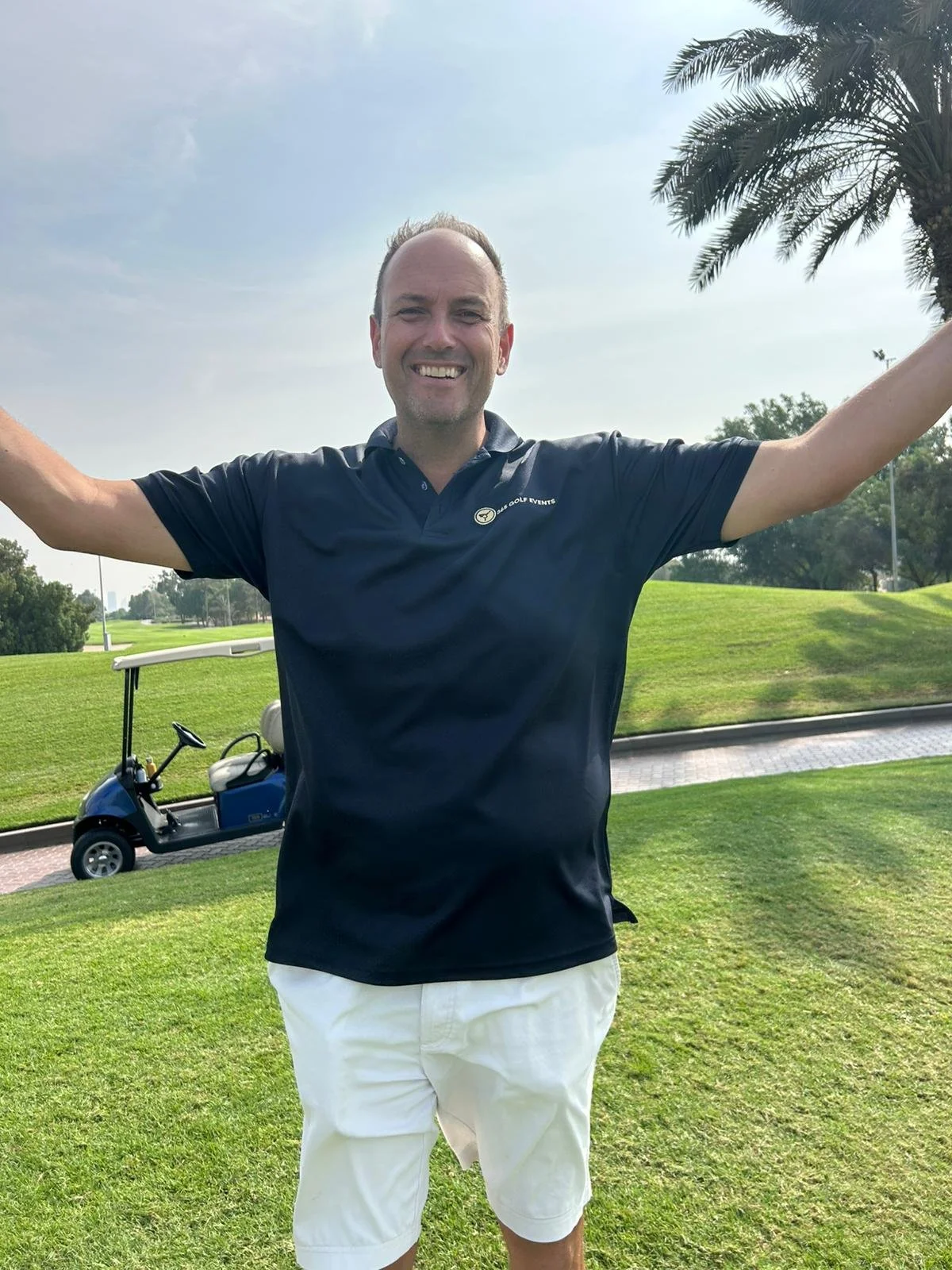 A man smiling with arms outstretched on a golf course, wearing a navy polo shirt and white shorts, with a golf cart in the background and a palm tree on the right.