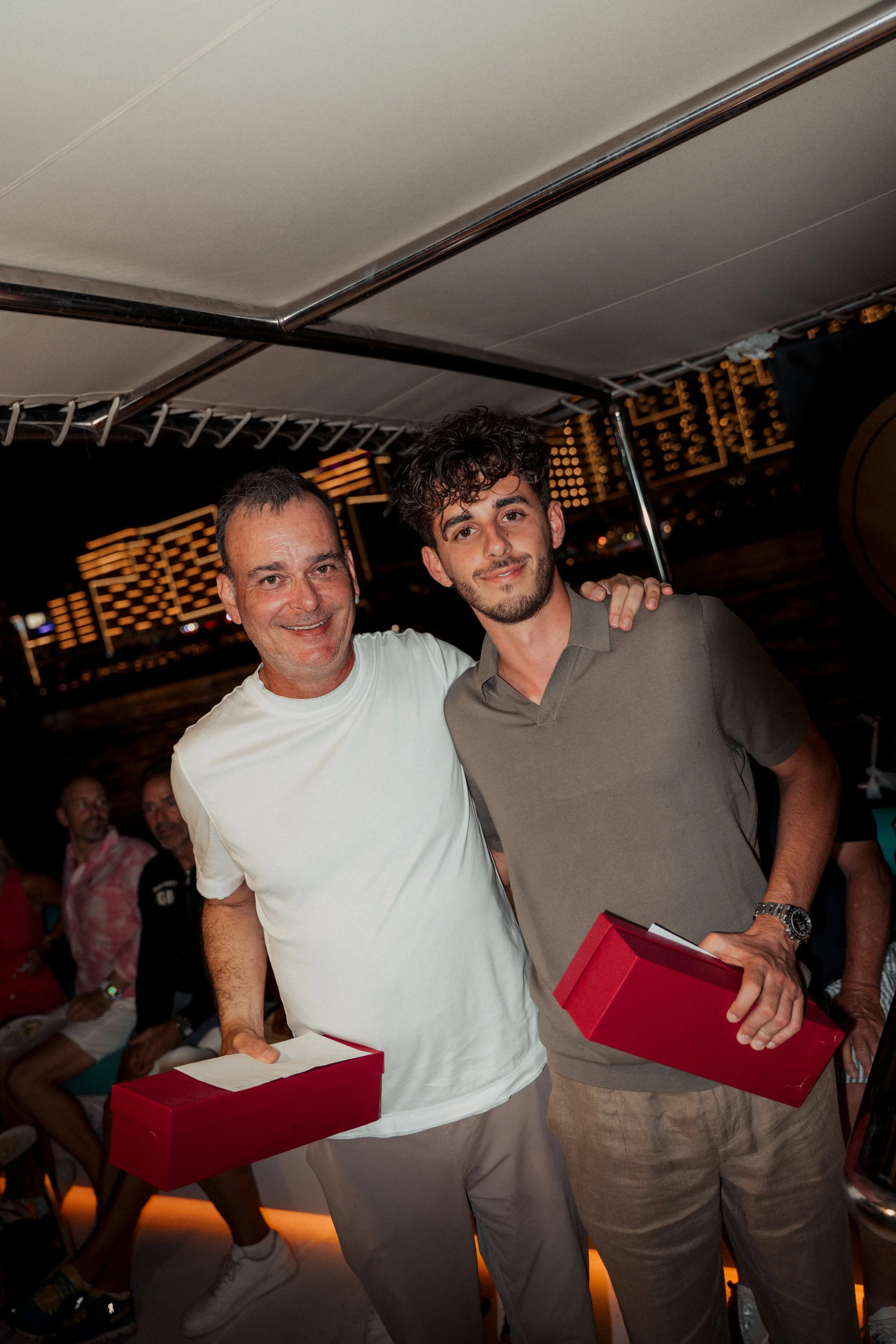 Two men smiling and holding red gift boxes, standing close together outdoors at night, with city lights and buildings in the background.