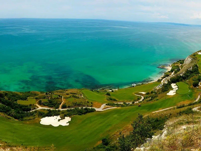 Aerial view of a golf course on a hillside overlooking the ocean with sand traps and green fairways