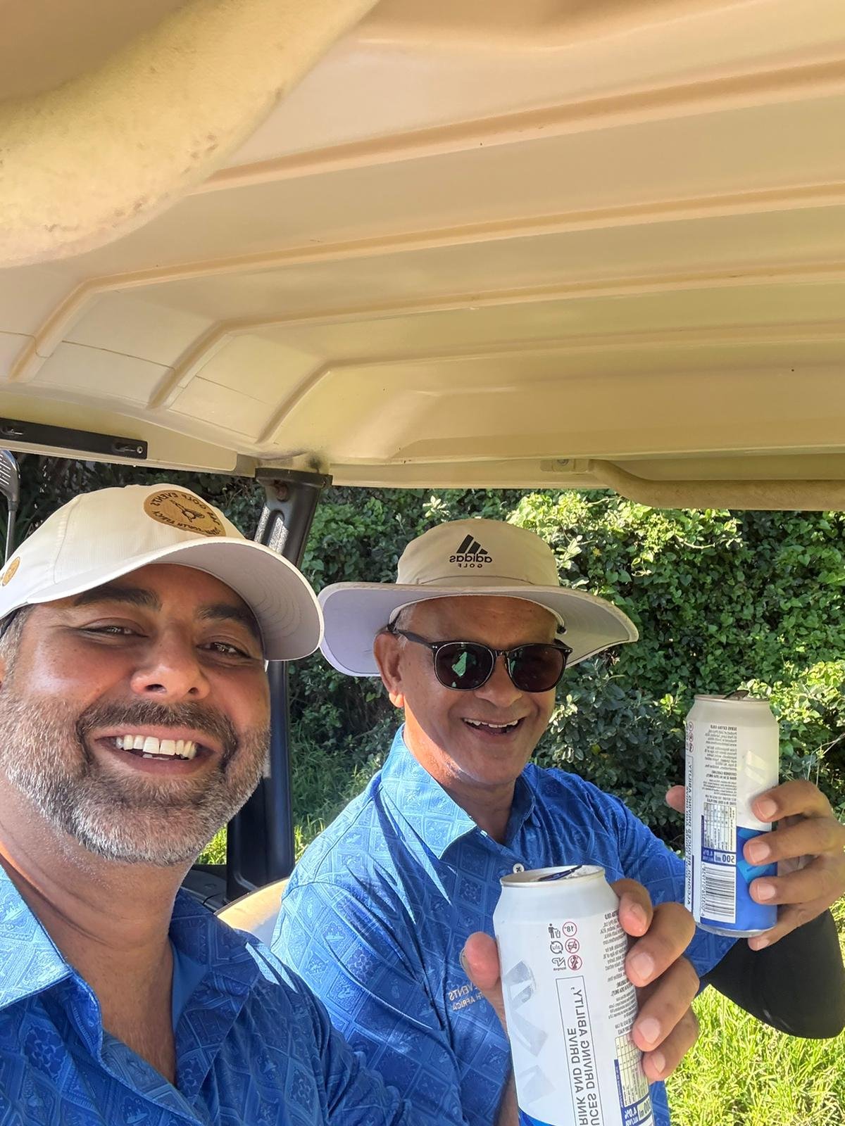Two men enjoying drinks outdoors next to a golf cart, wearing hats and sunglasses, with greenery in the background.