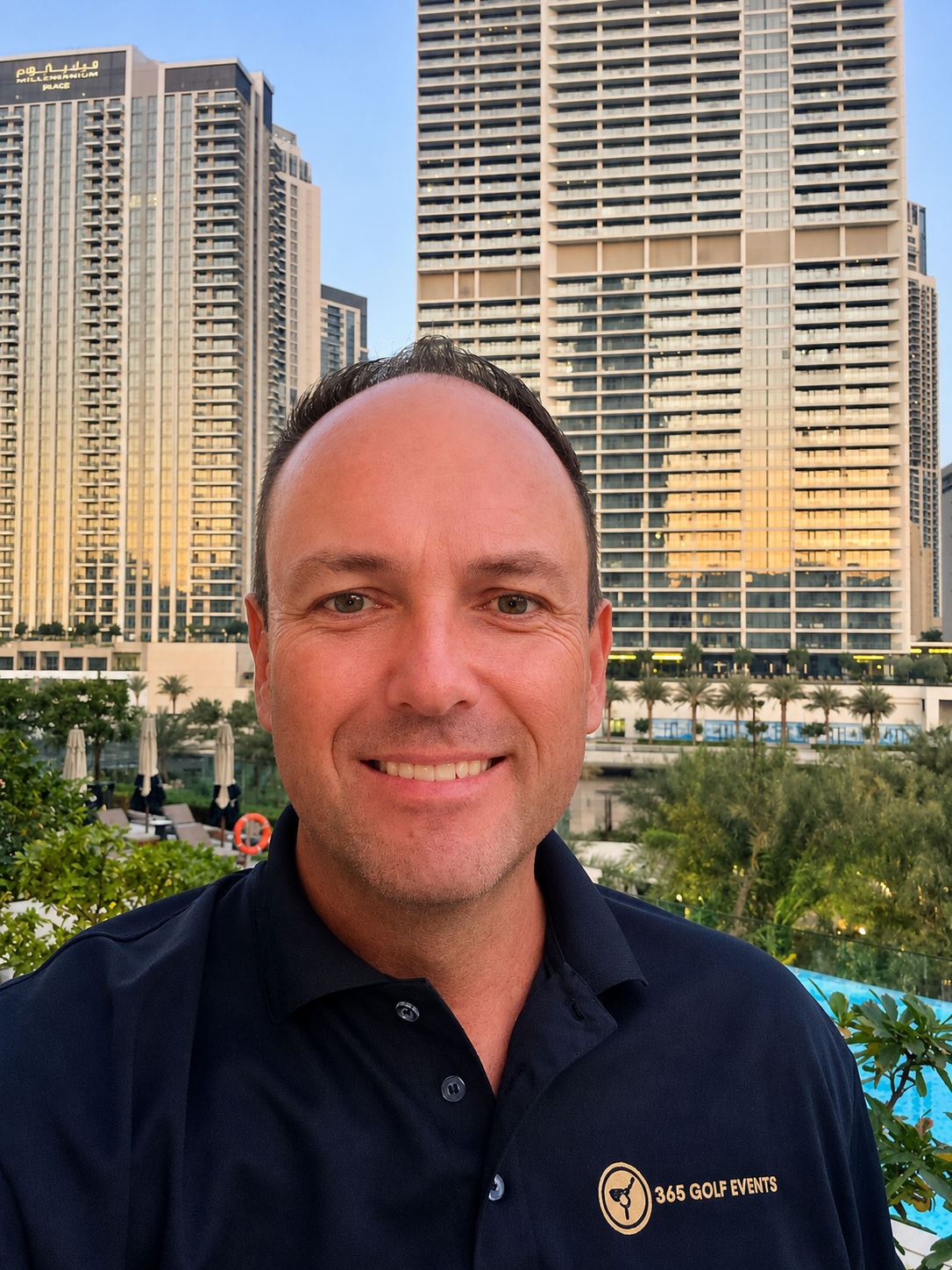 A man smiling outdoors in front of tall, modern skyscrapers during sunset or early evening, with a lush green environment and a swimming pool area nearby.