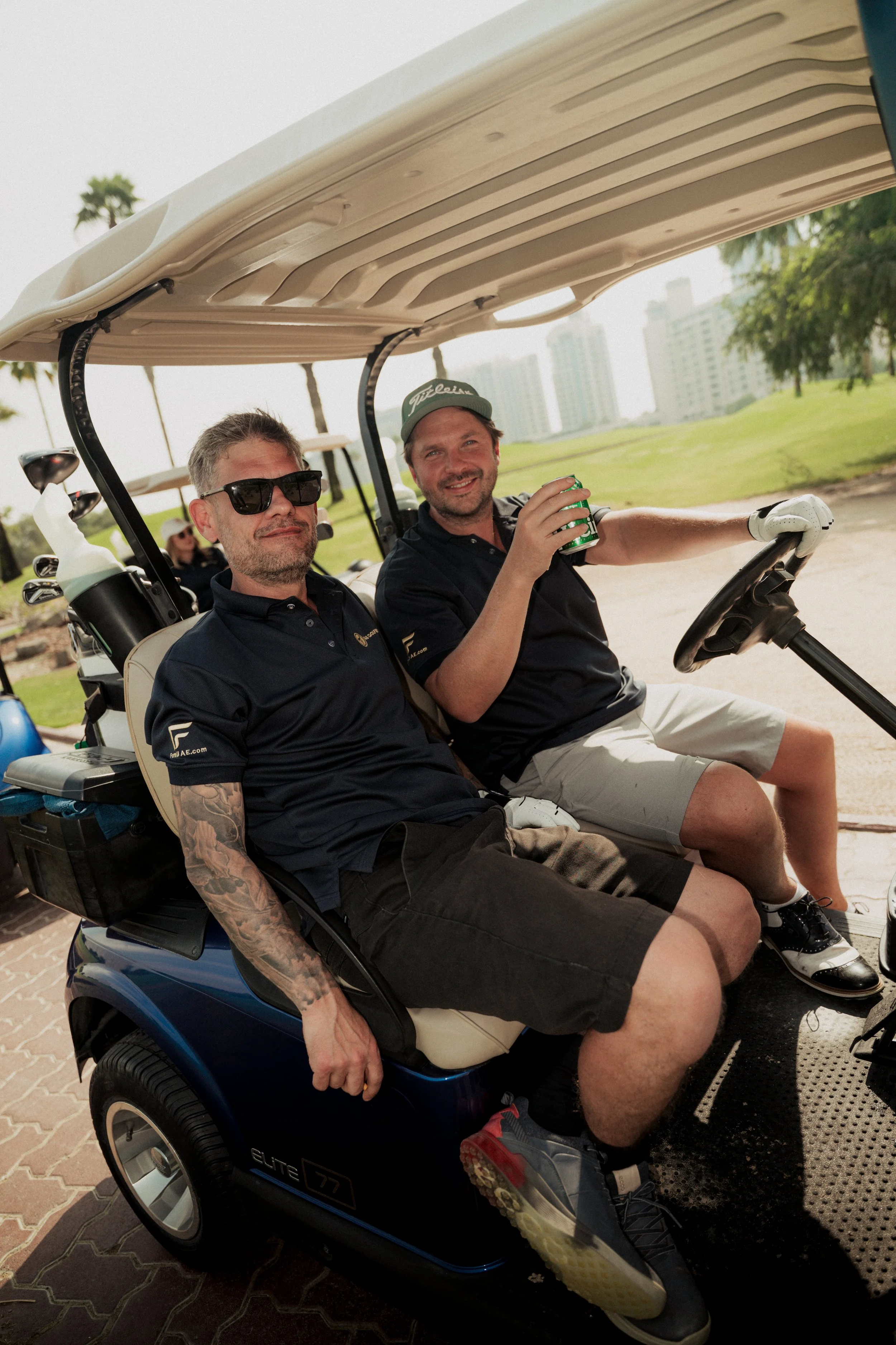 Two men sitting in a golf cart on a golf course, smiling and holding drinks. One man is wearing sunglasses, the other is wearing a cap. They are surrounded by trees and tall buildings in the background.