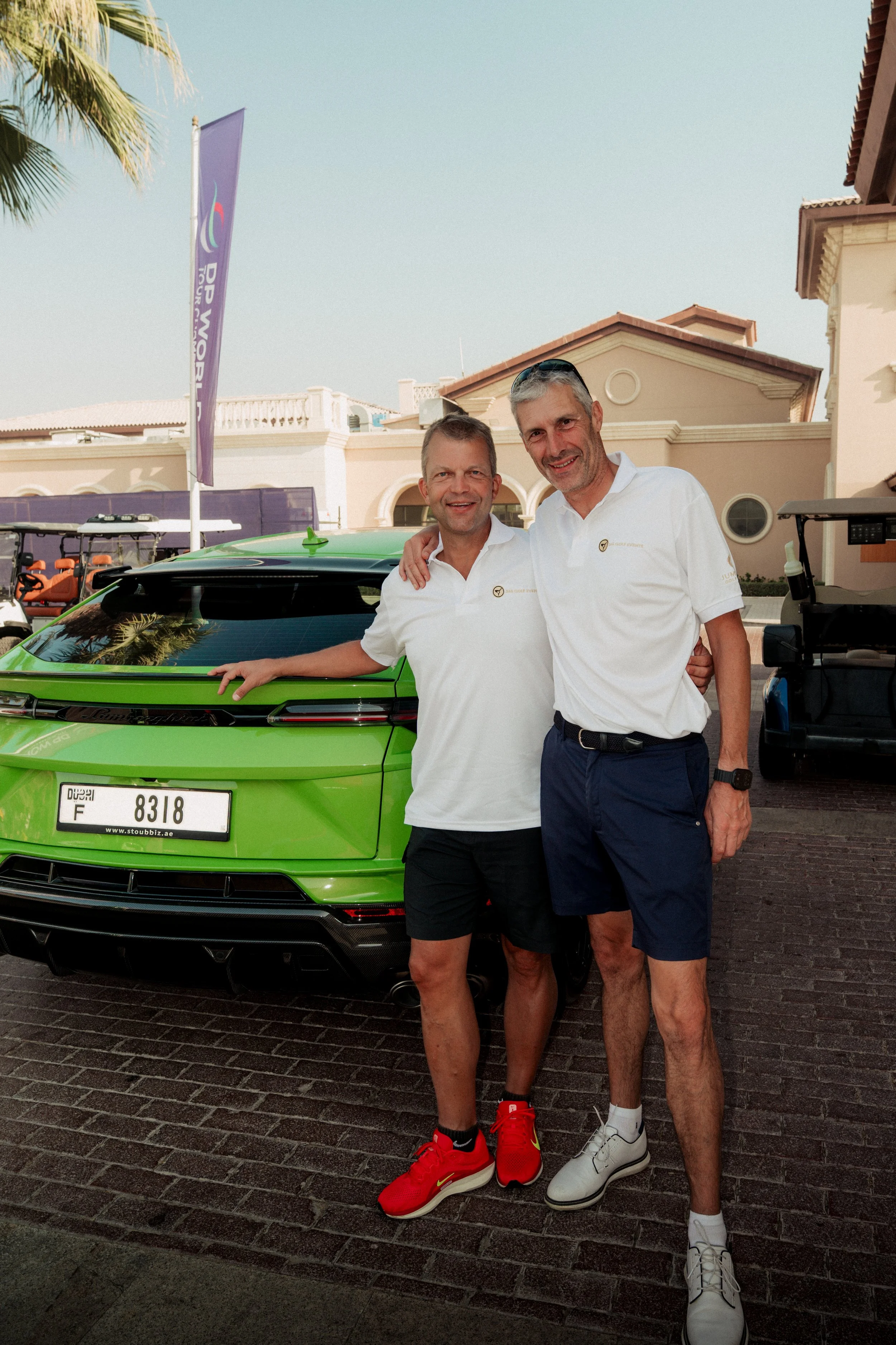 Two men standing beside a bright green sports car, smiling, with one arm around each other, in a sunny outdoor setting near a building, with a golf cart in the background.