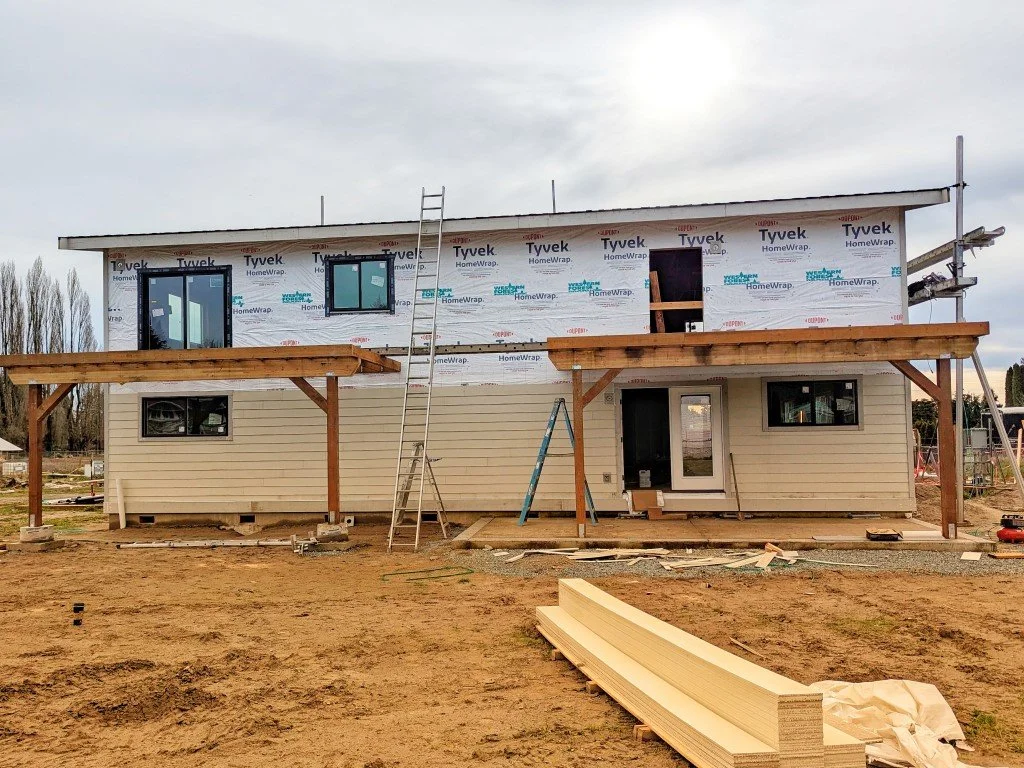 Backside of current SFR newbuild project. across the flattened earth home has half the siding installed with two wooden gondolas.