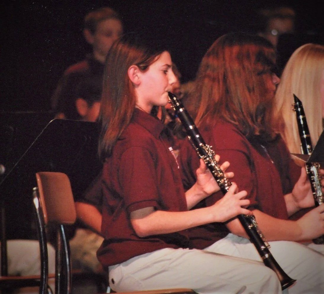 A group of tweens playing clarinets in a performance, seated in a row, wearing matching maroon shirts, with some reading sheet music, and other players visible in the background.