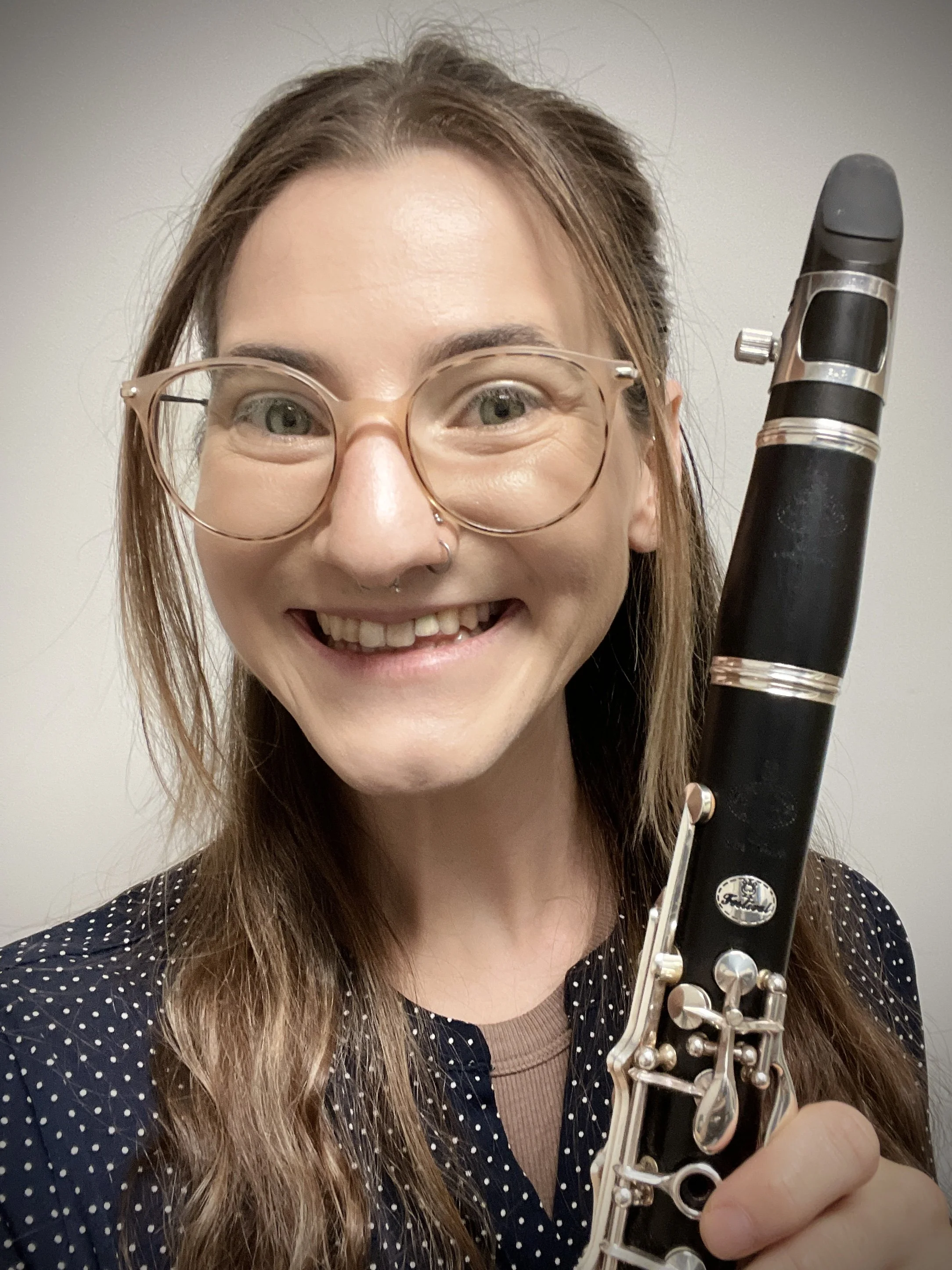 A woman with glasses smiling while holding a clarinet in front of a plain background.