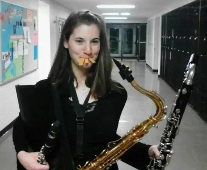 Young woman holding a saxophone and a clarinet in a school hallway, with reeds her mouth, giving a playful expression.