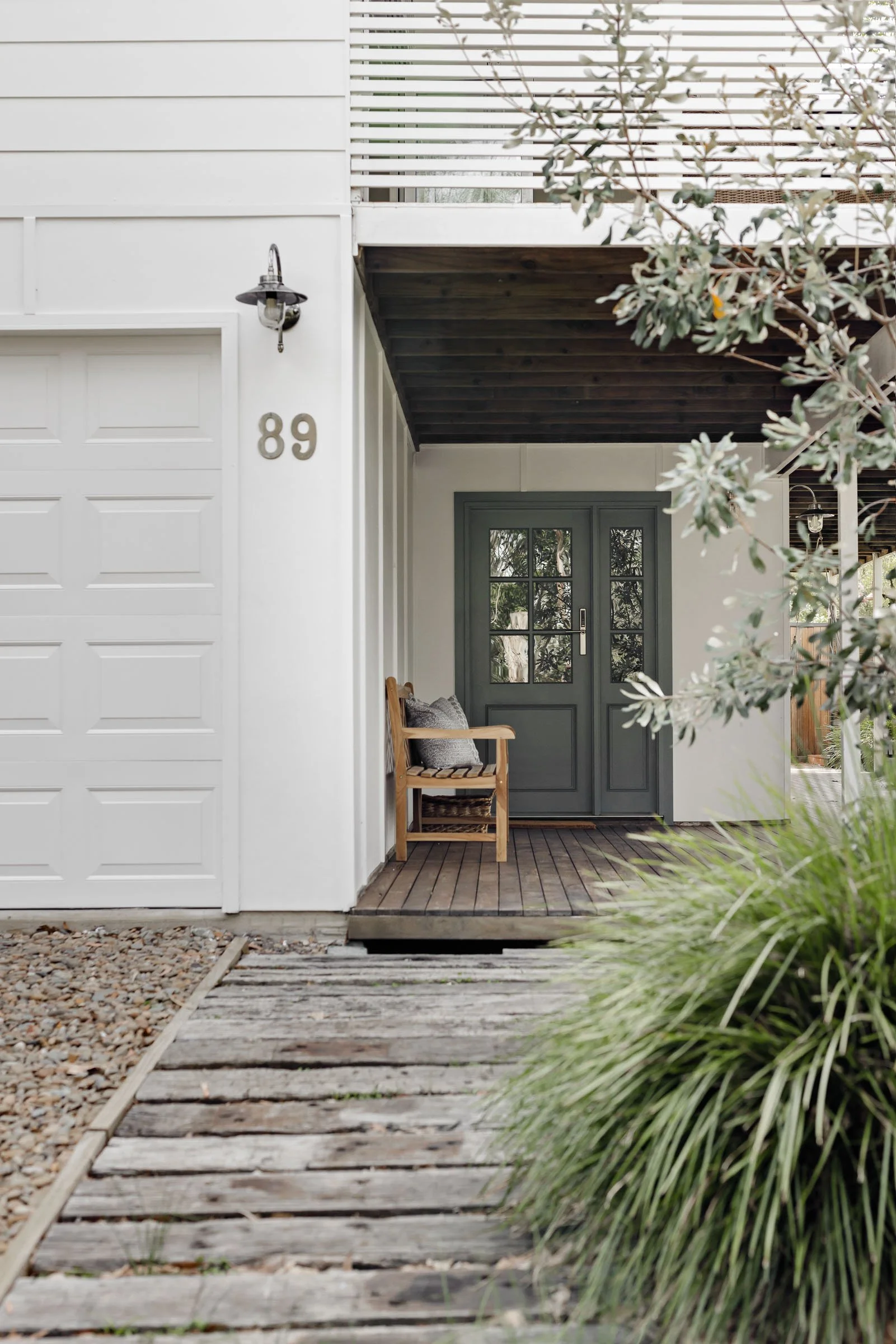 Front entrance of a modern house with a gray door, a wooden bench with pillows, a white garage door, house number 89, outdoor lighting, and a wooden path leading to the door surrounded by greenery.