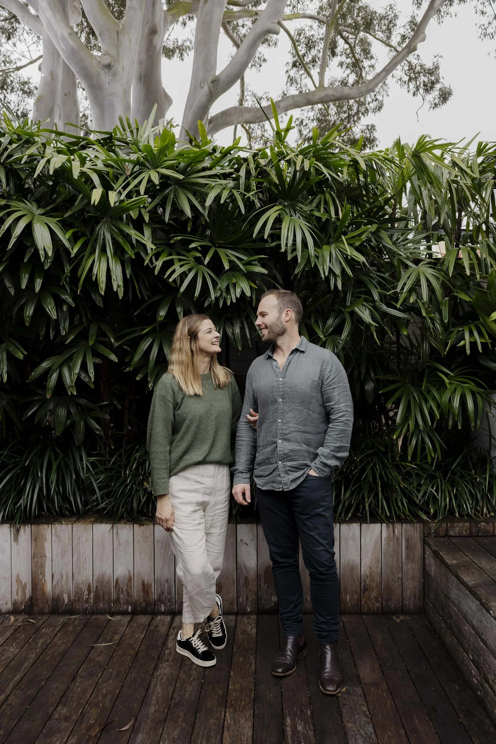 A woman and man standing on wooden deck in front of large green foliage, smiling at each other, woman in green sweater and beige pants, man in gray shirt and dark pants.