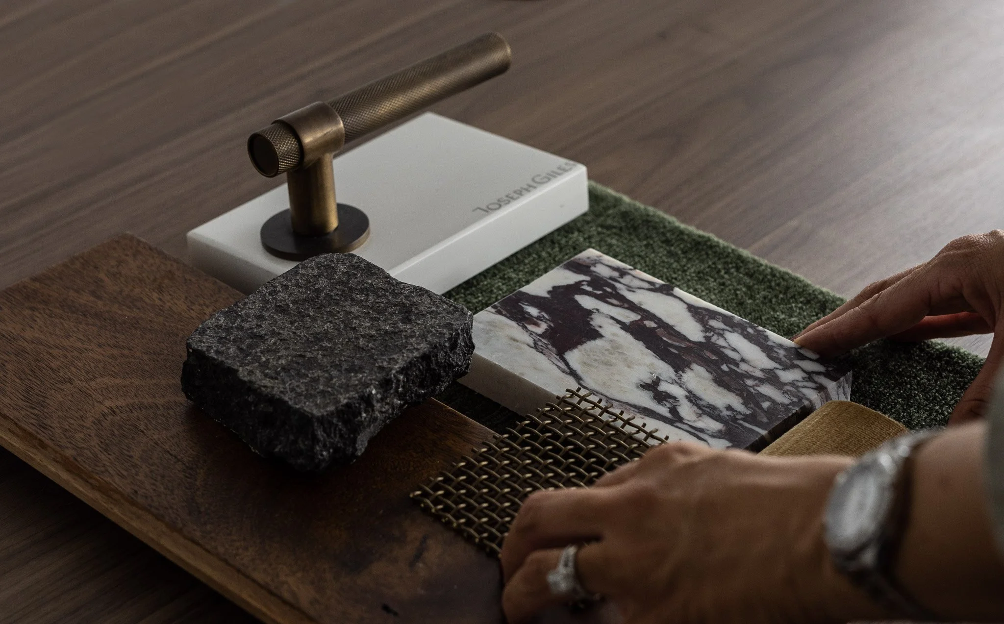 Person examining a marble tile on a wooden table surrounded by a black stone slab, a small metal mesh, and a sample with a white and black marble pattern.