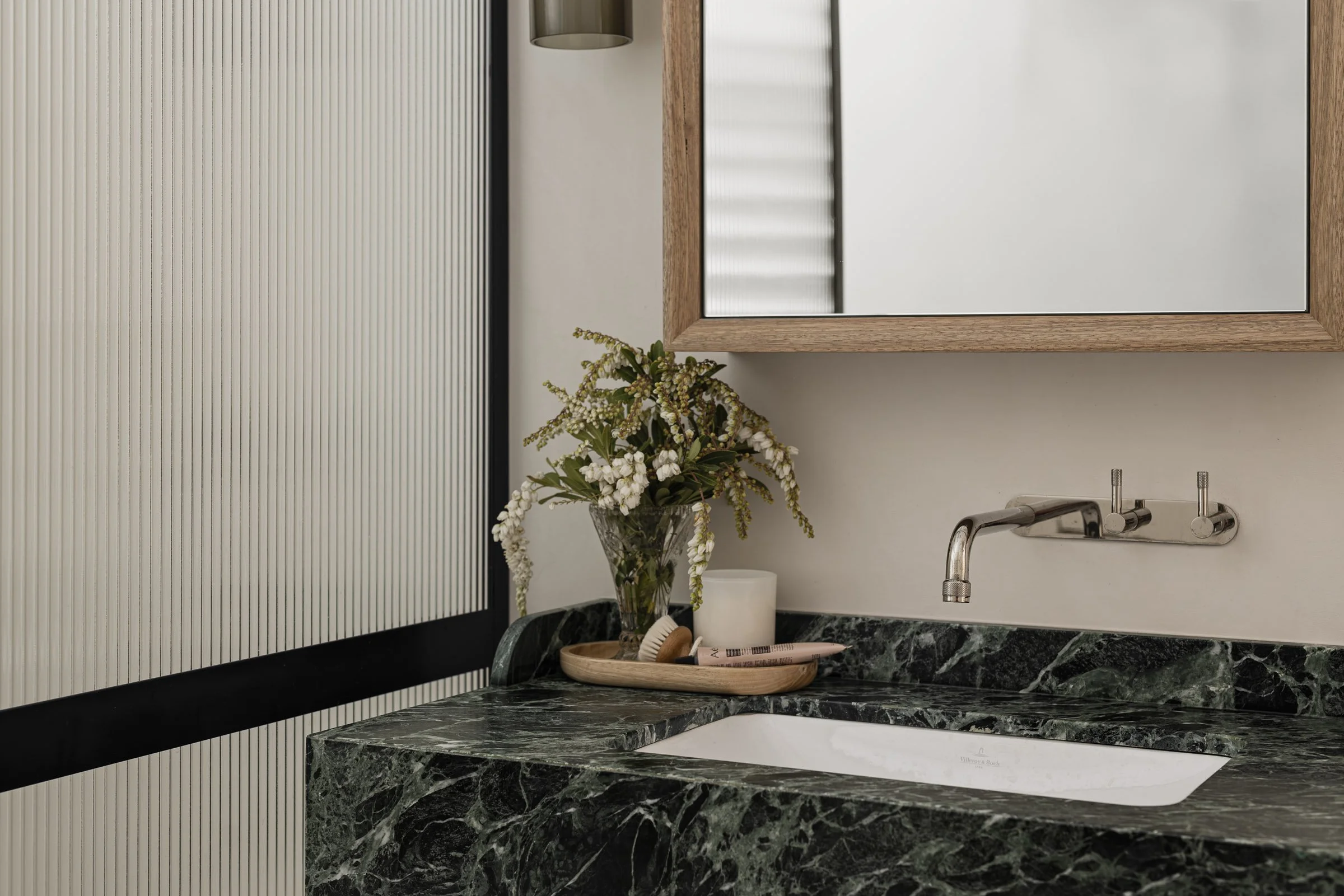 Bathroom sink with a black marble countertop, a wooden tray with toiletries, a green vase with white flowers, and a large mirror with a wooden frame.