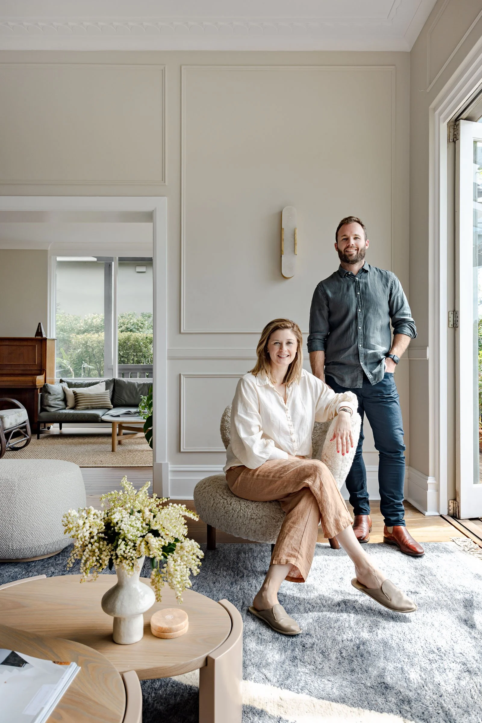 A smiling woman sitting in a chair and a smiling man standing next to her in a well-lit, modern living room.