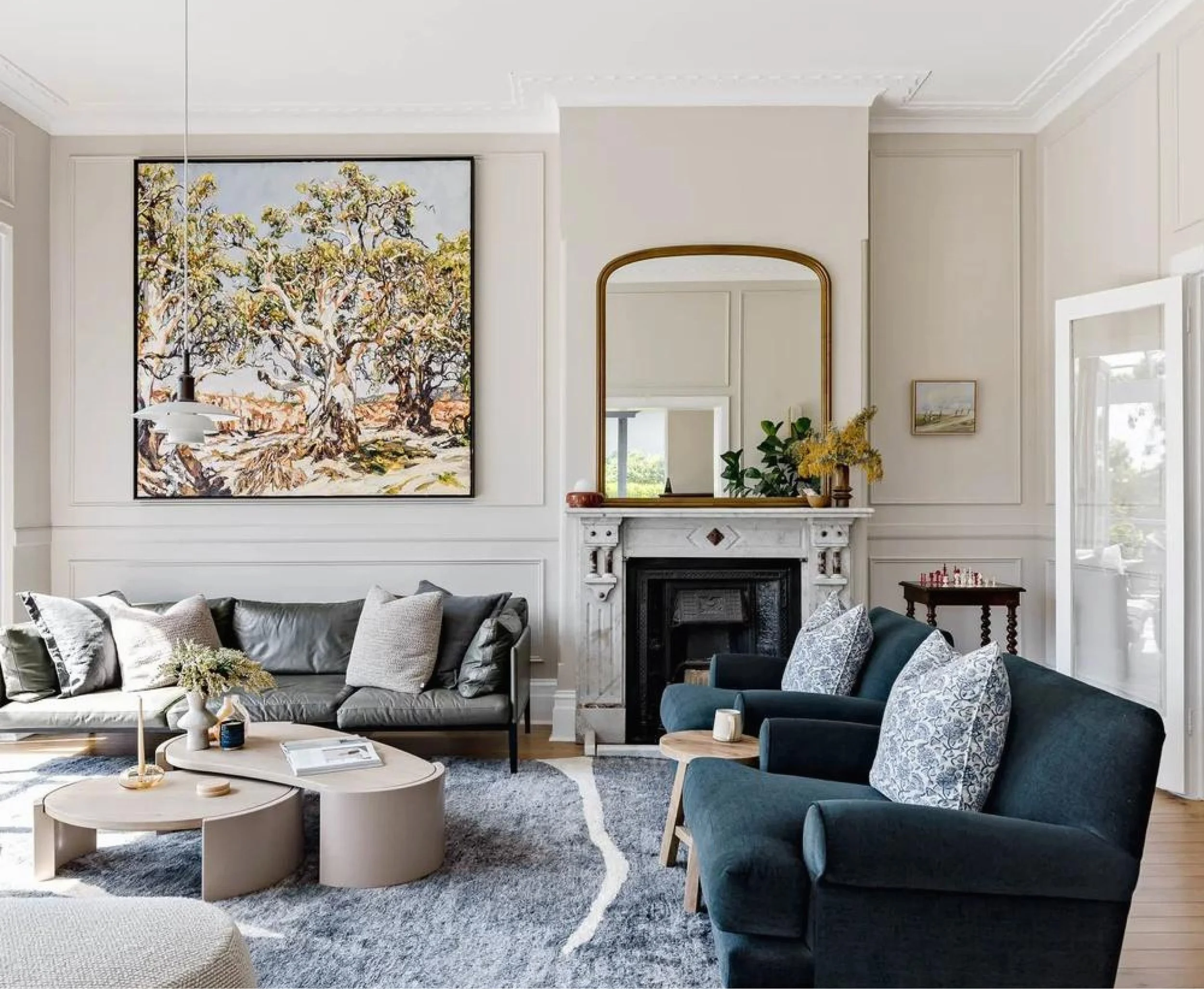 Living room with a large framed tree artwork above a gray leather sofa, a fireplace with a gold-framed mirror, dark blue armchair with patterned pillows, marble fireplace mantel decorated with a plant and a small vase, coffee table, and a chess set in the background.