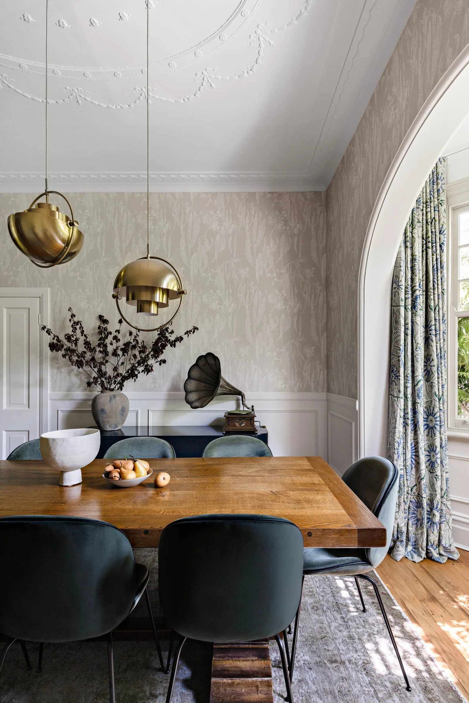 A dining room with a large wooden table, five green chairs, a vintage gramophone, a vase with dried flowers, and a bowl of pears. The room has light-colored wallpaper, decorative ceiling molding, and large window with floral-patterned curtains. Two gold pendant lights hang from the ceiling.