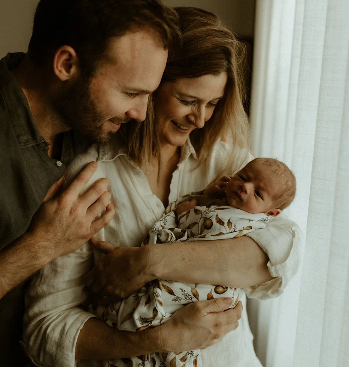 A woman holding a newborn baby near a window, with a man leaning in to look and smile at the baby.