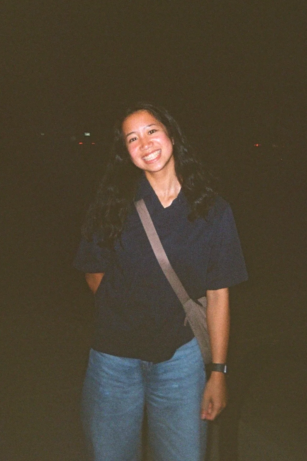 A young woman with long dark hair, smiling, standing outdoors at night in a dark shirt and light blue jeans, with a beige crossbody bag.