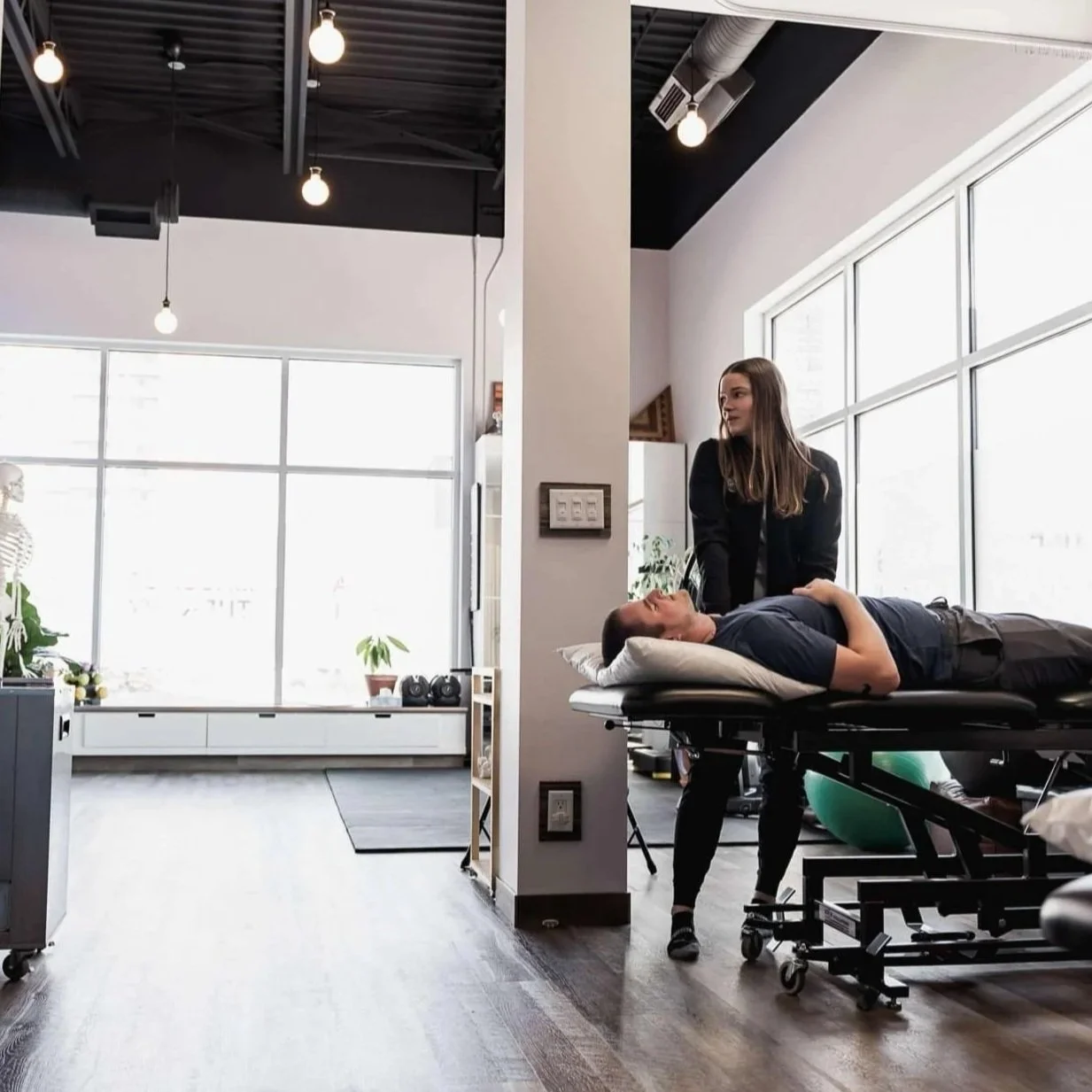Athletic therapist providing hands-on treatment in a Winnipeg injury and pain rehabilitation clinic on a male patient lying on a treatment table in a modern, well-lit office with large windows, plants, and therapy equipment.