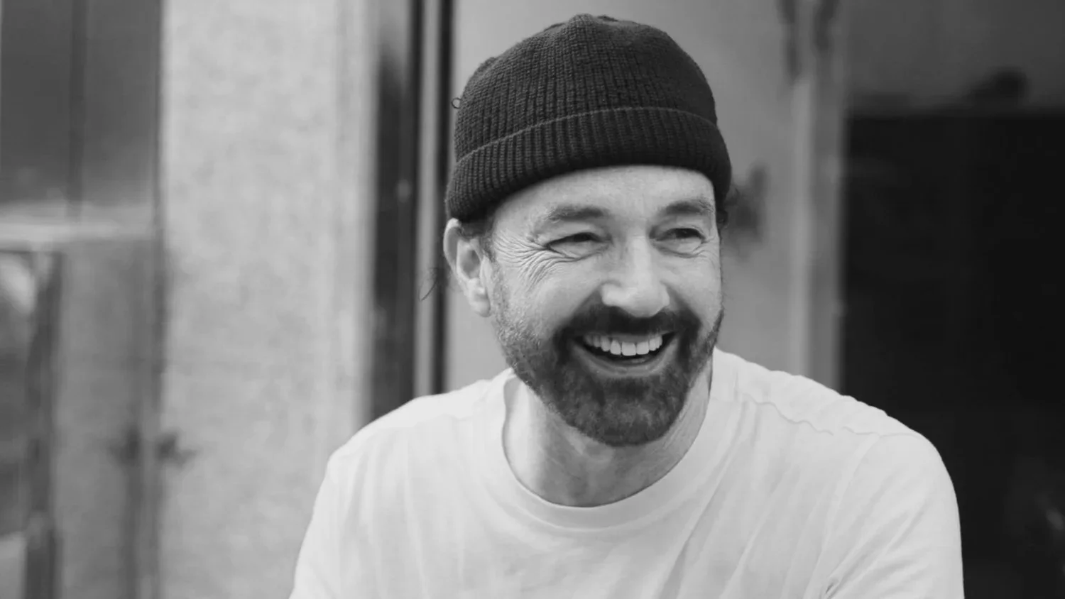 A man with a beard and mustache, wearing a knit cap and a white shirt, smiling and appearing happy.