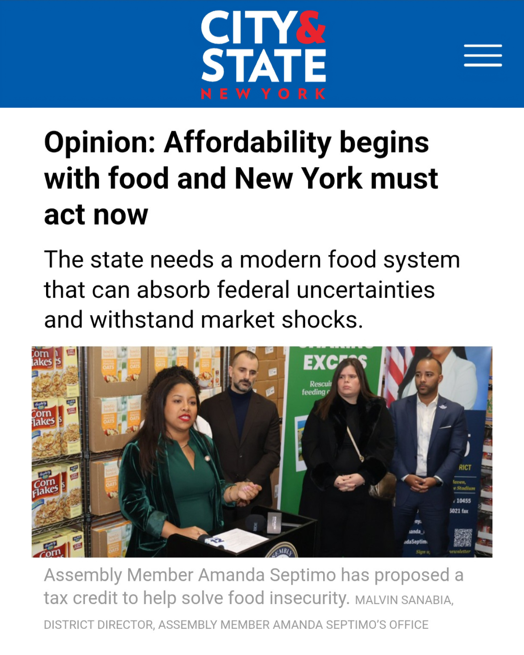 A woman speaking at a press conference with a microphone, surrounded by other people, in front of shelves stacked with cereal boxes and a green banner.